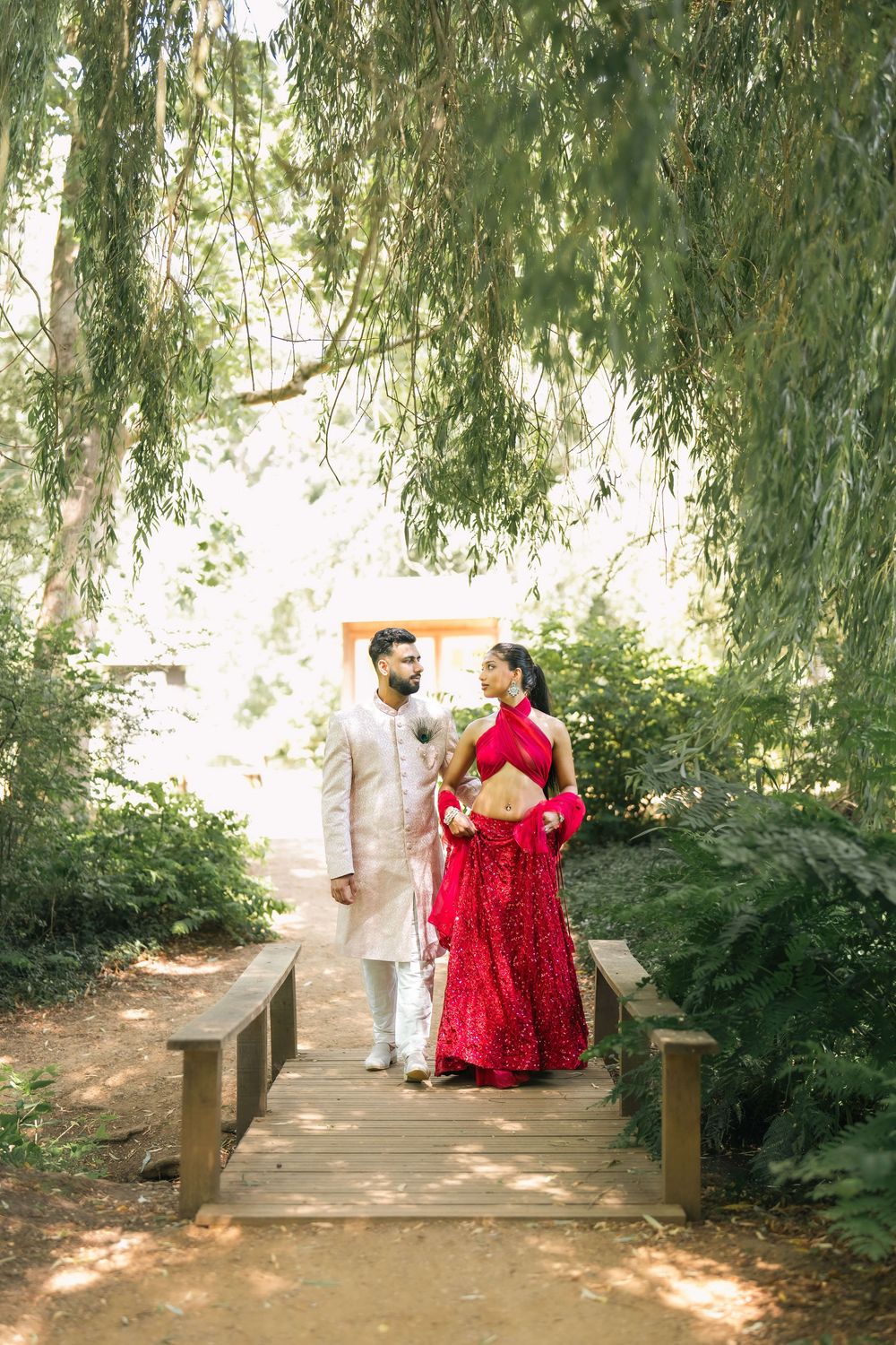 Indian couple in traditional wedding attire standing on a wooden bridge surrounded by lush green trees.