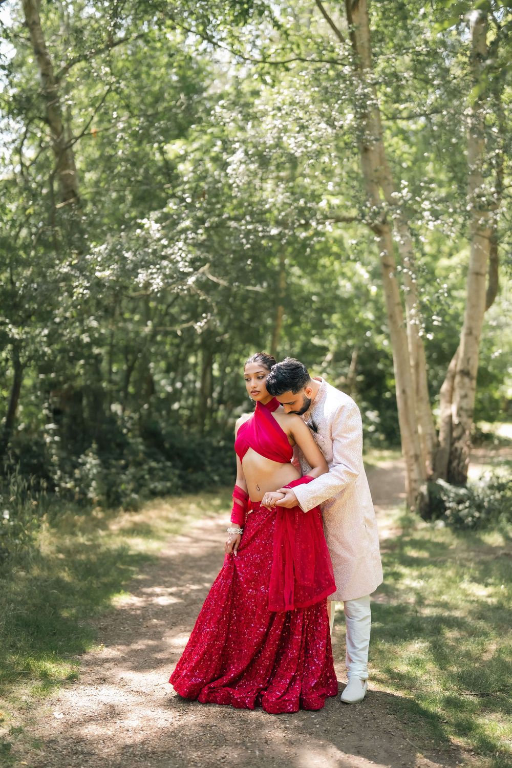 Couple in traditional Indian wedding attire embracing on a forest path, bride in red lehenga, groom in white sherwani.
