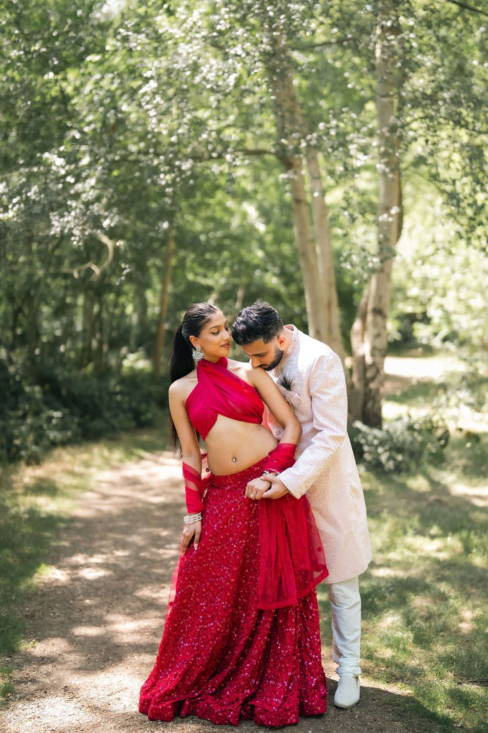 Couple in traditional Indian attire embracing outdoors, woman in red lehenga, man in white sherwani, lush green trees.