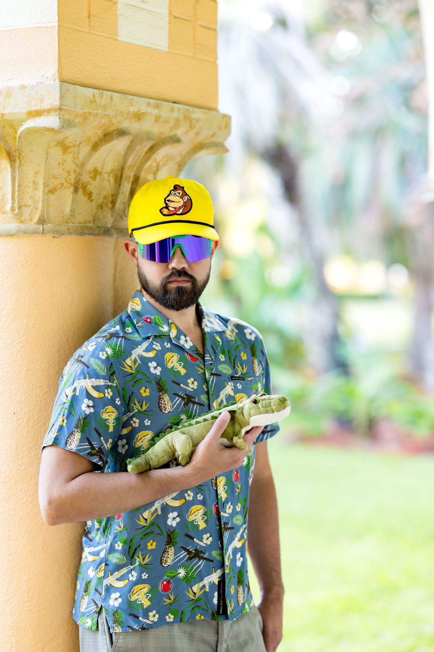 Bearded man in tropical shirt, yellow cap, and mirrored sunglasses holding green vegetable by ornate column outdoors.