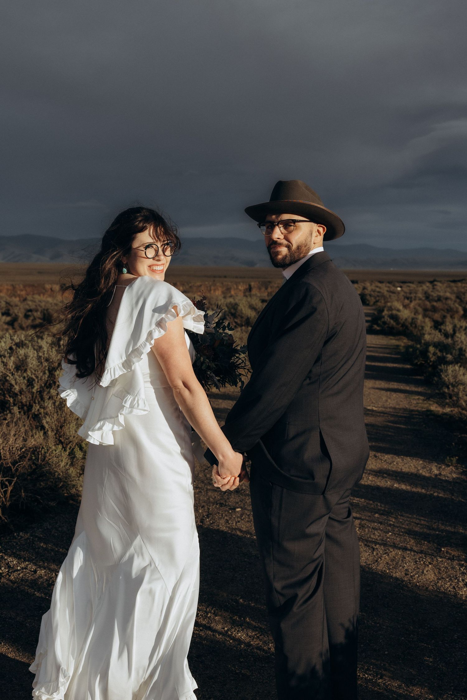 A couple in wedding attire holds hands in a desert landscape, bride in white ruffled gown, groom in dark suit and hat.
