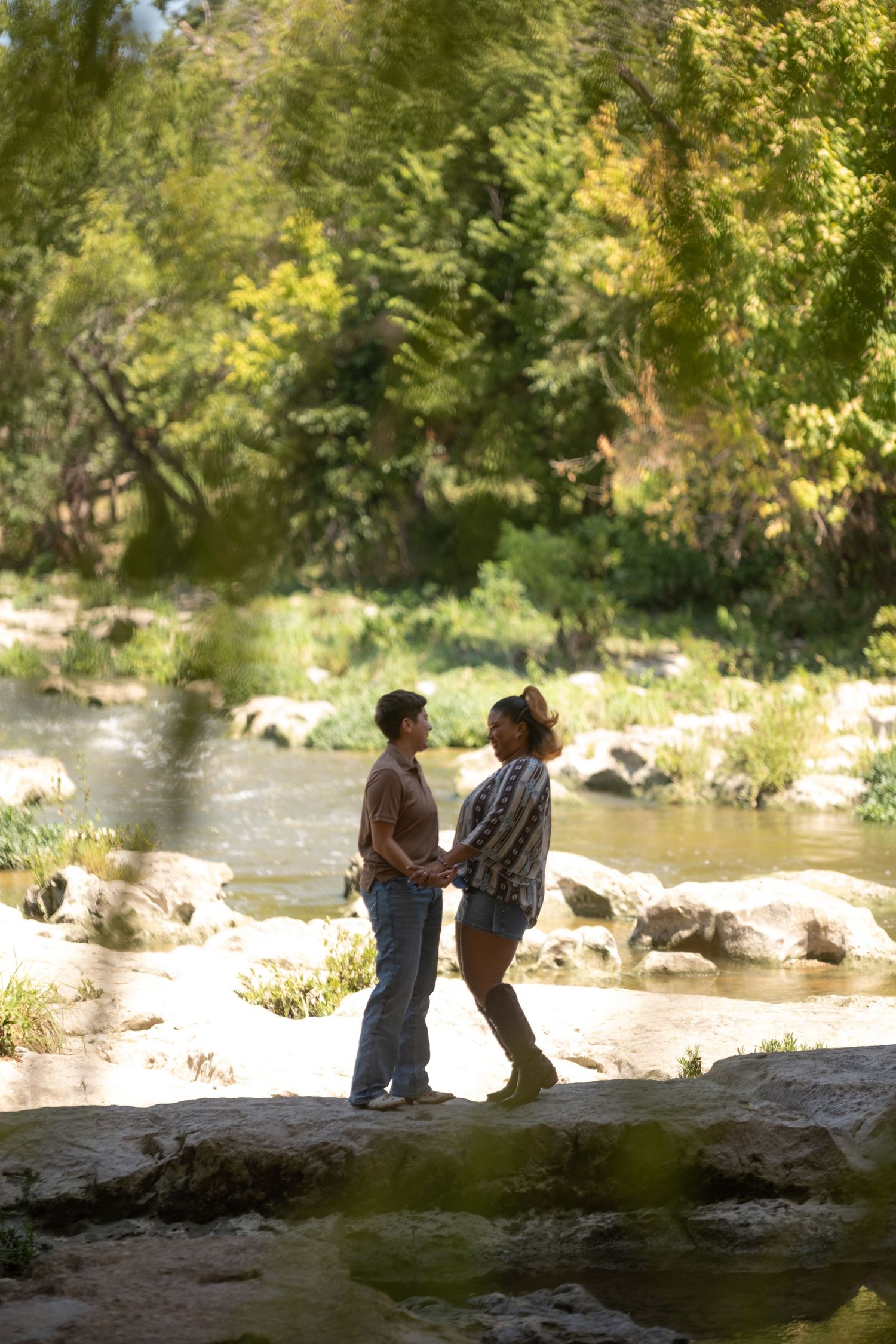 Couple holding hands by a rocky stream surrounded by lush green trees in a romantic outdoor setting.