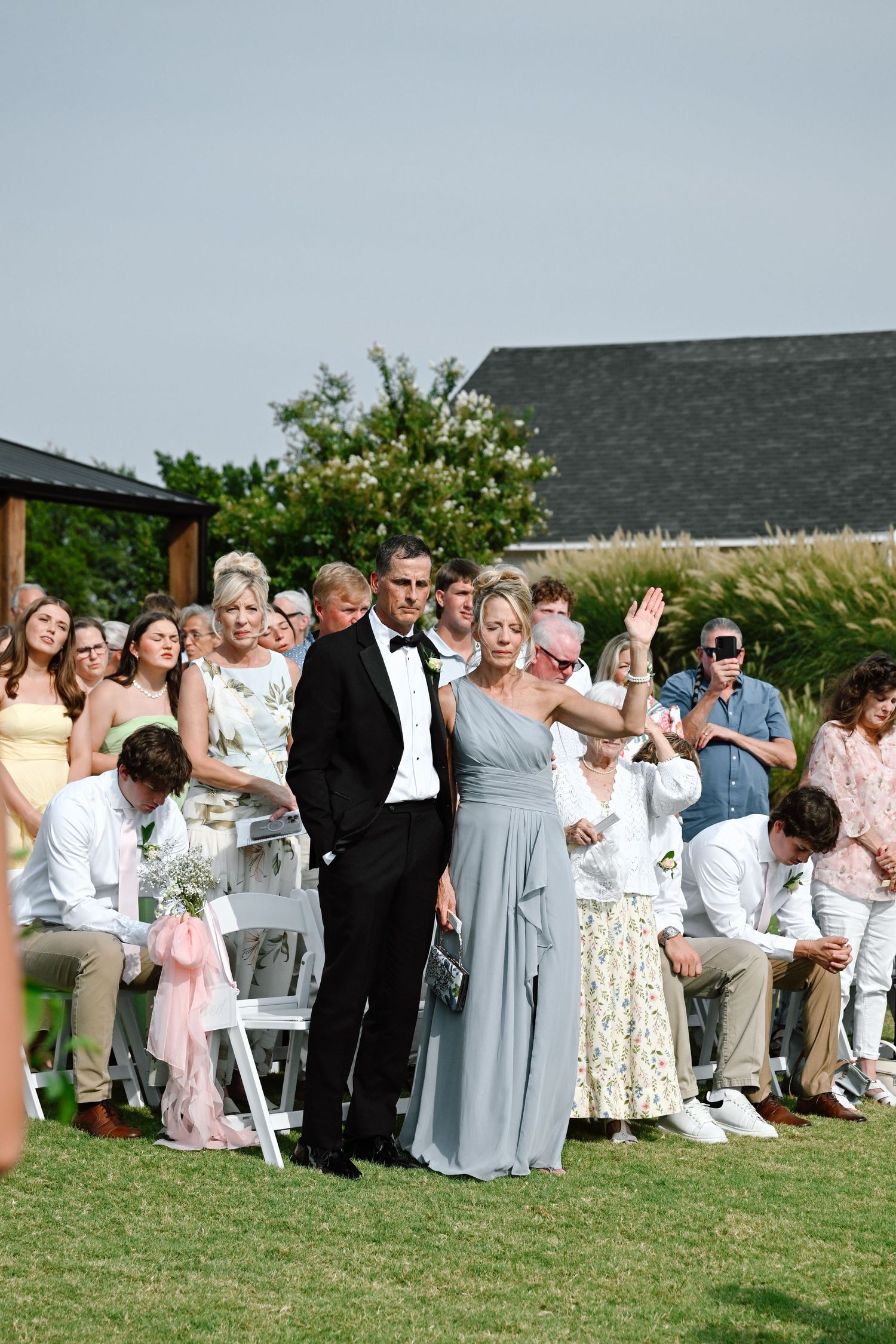 Outdoor wedding ceremony with guests seated on white chairs, a couple walks down the aisle on a sunny day.