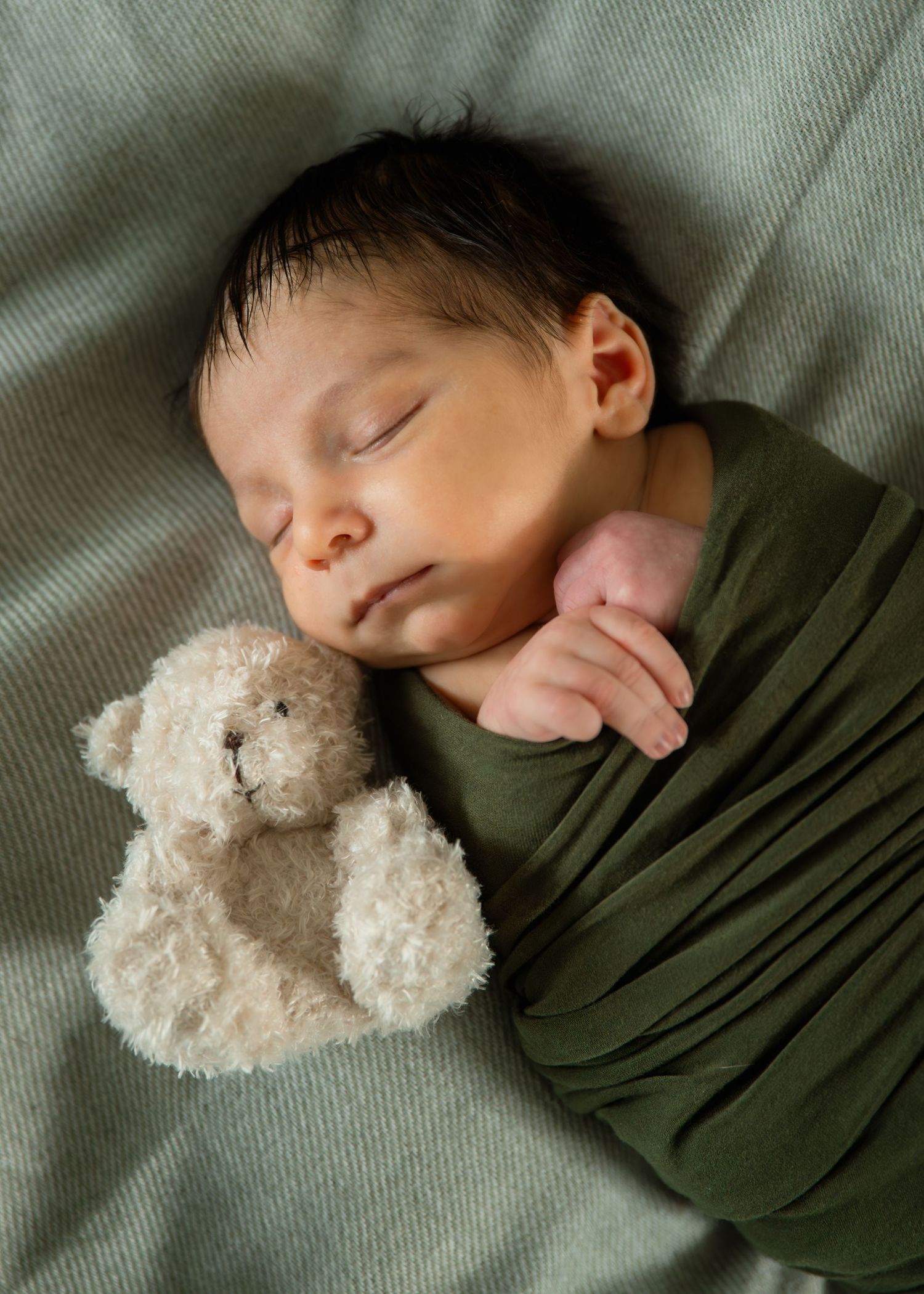 Sleeping newborn baby swaddled in green wrap cuddles a small teddy bear on a soft gray blanket.