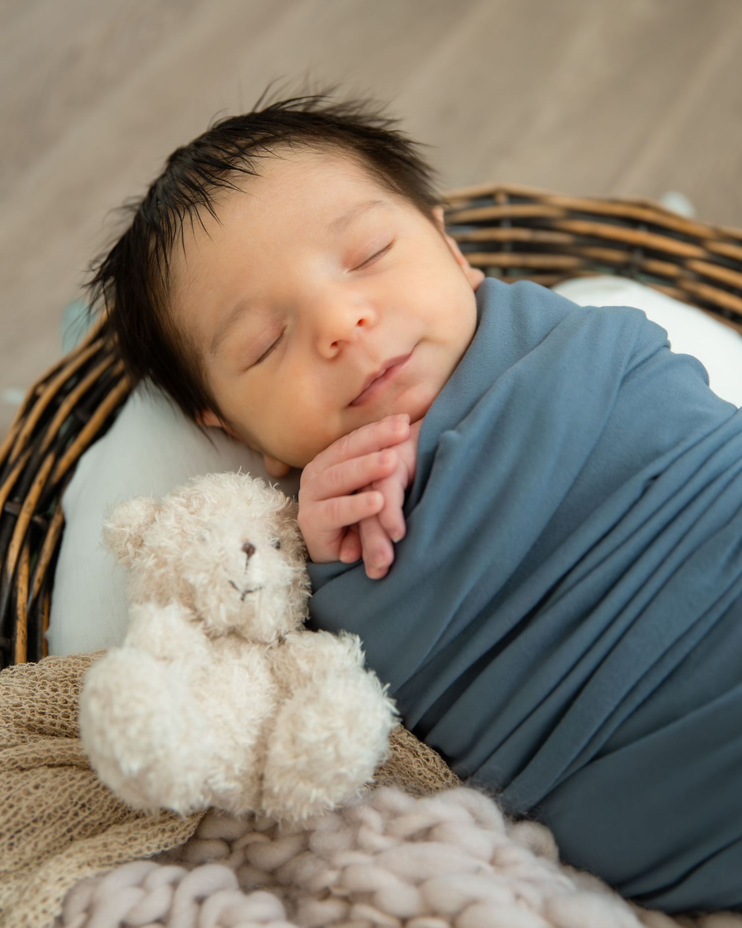 Sleeping newborn baby swaddled in blue wrap cuddles a tiny teddy bear inside a wicker basket.