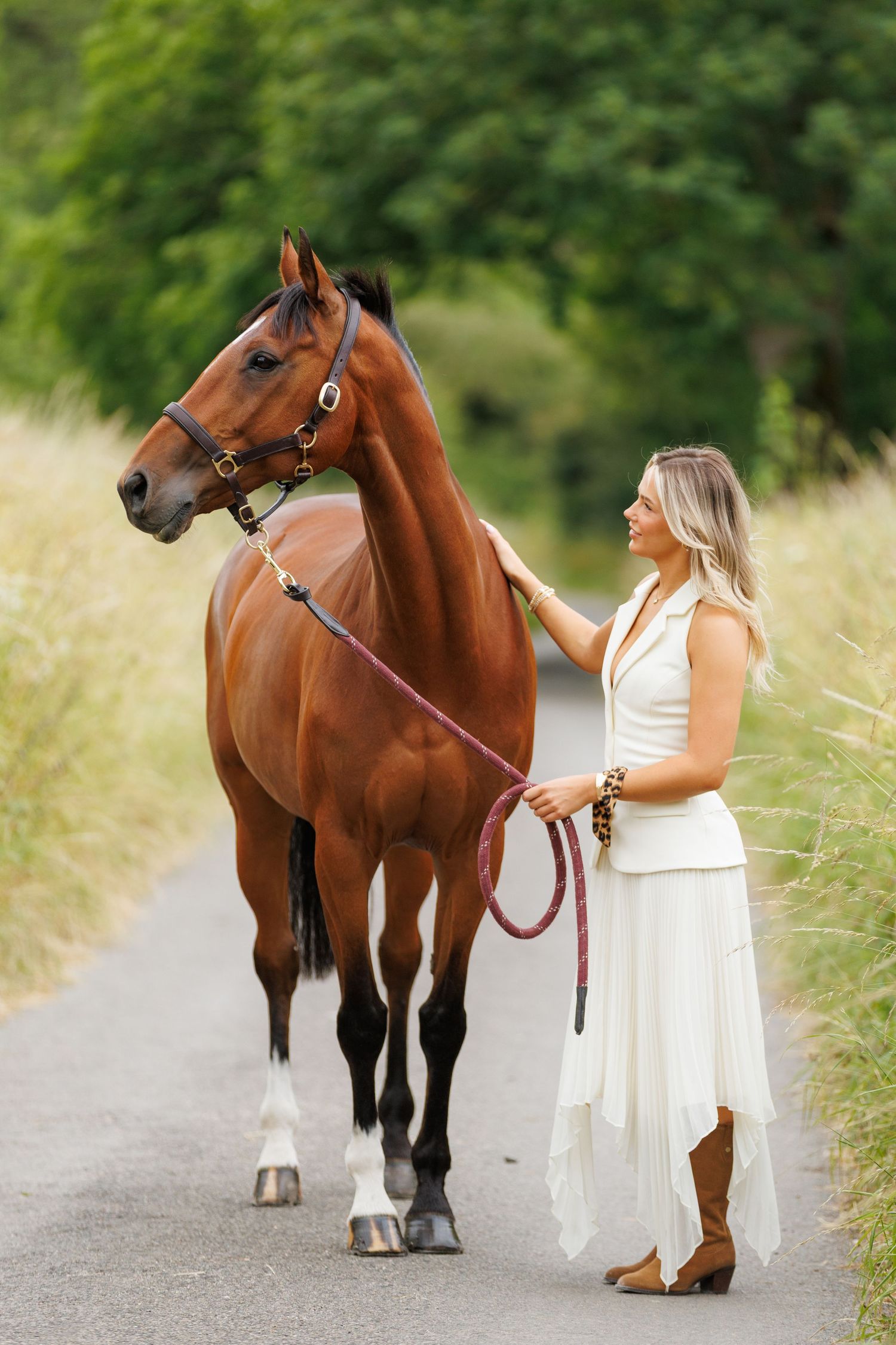 A beautiful bay horse wearing a black bridle stands next to someone in a white lace dress in an outdoor setting.