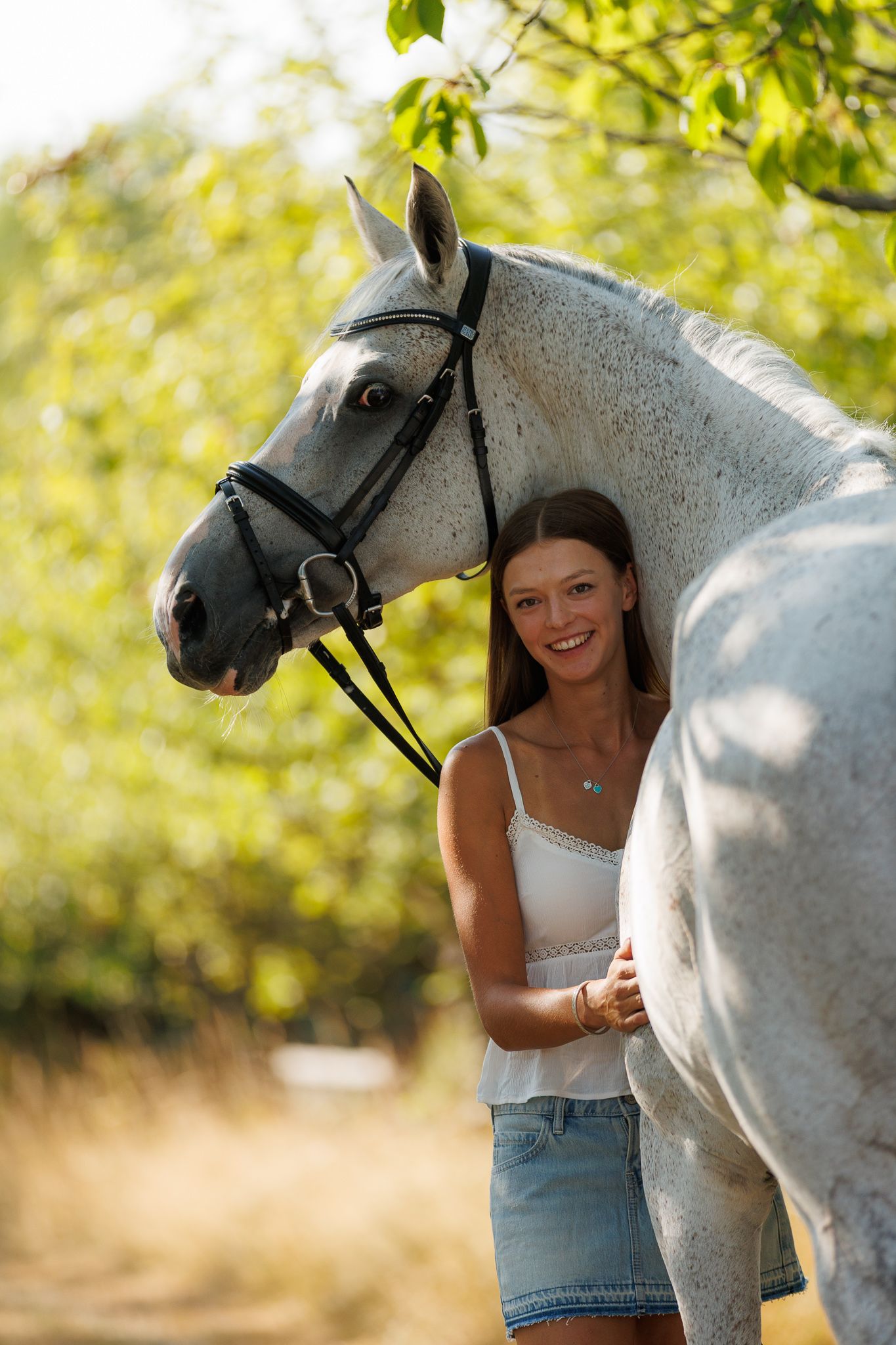 Young woman smiling between two bay horses with white markings, holding reins near a brick stable.