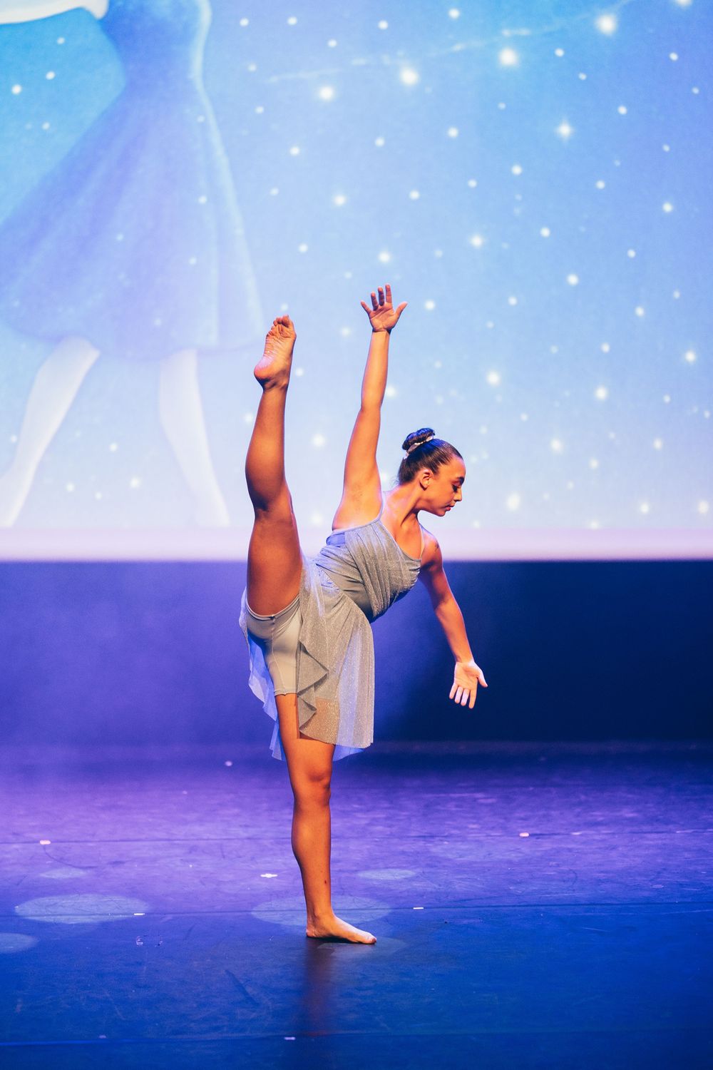 A young dancer performs an elegant arabesque on stage in a gray costume under blue stage lighting.