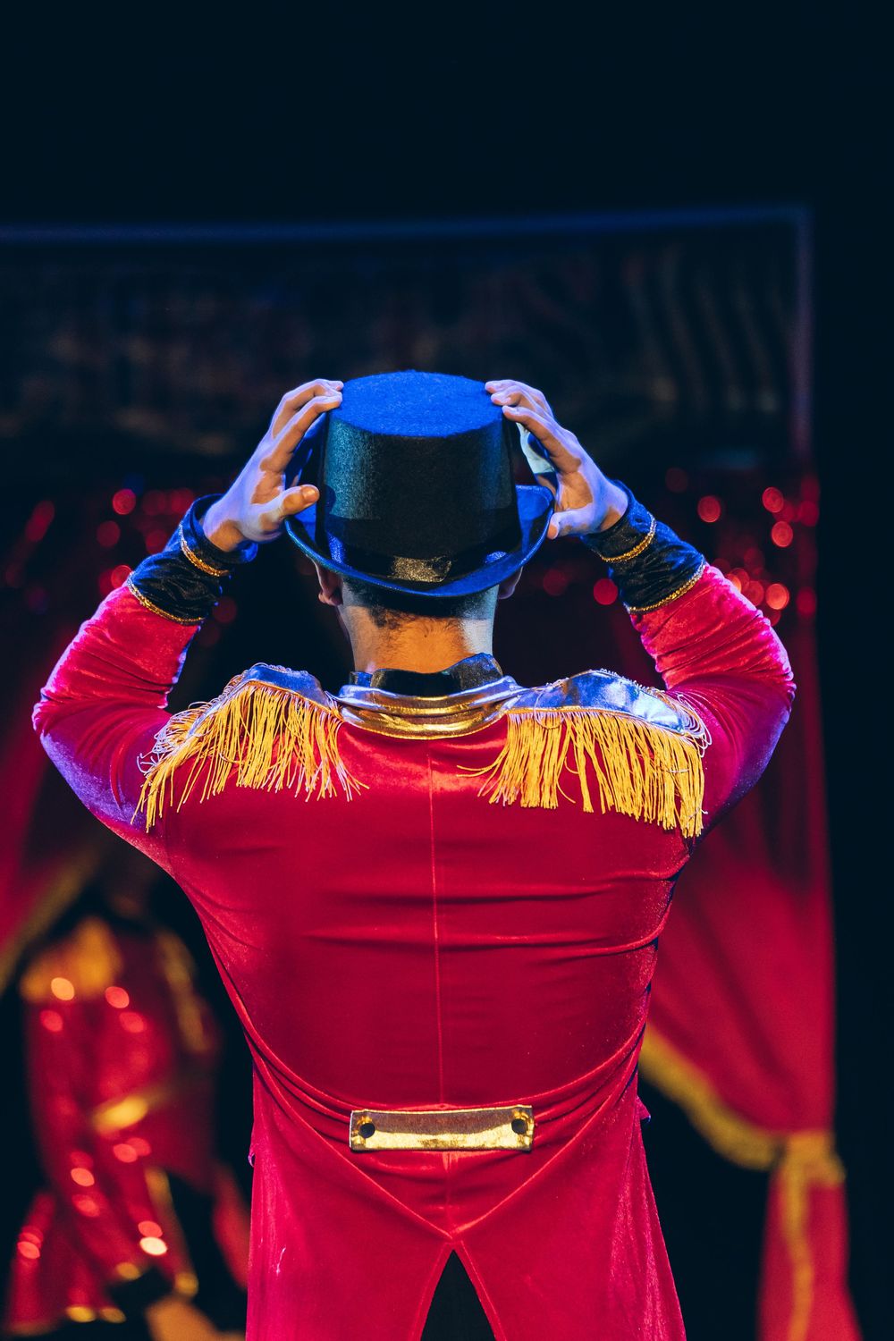 Circus ringmaster in red velvet costume with gold epaulettes adjusts black top hat on stage.