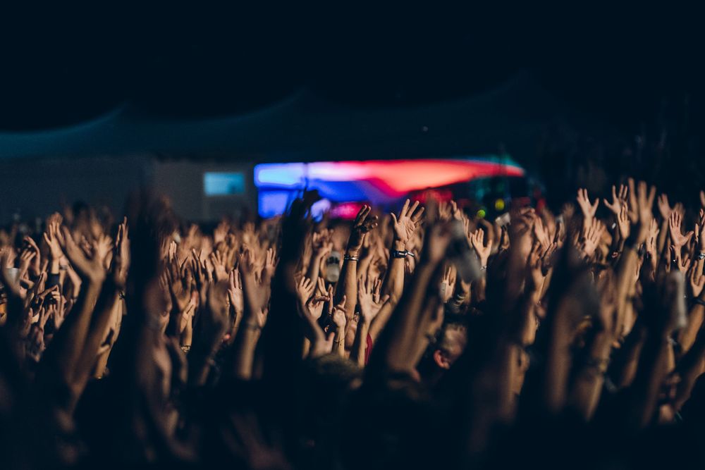 A sea of raised hands at a concert with colorful stage lighting in the background.