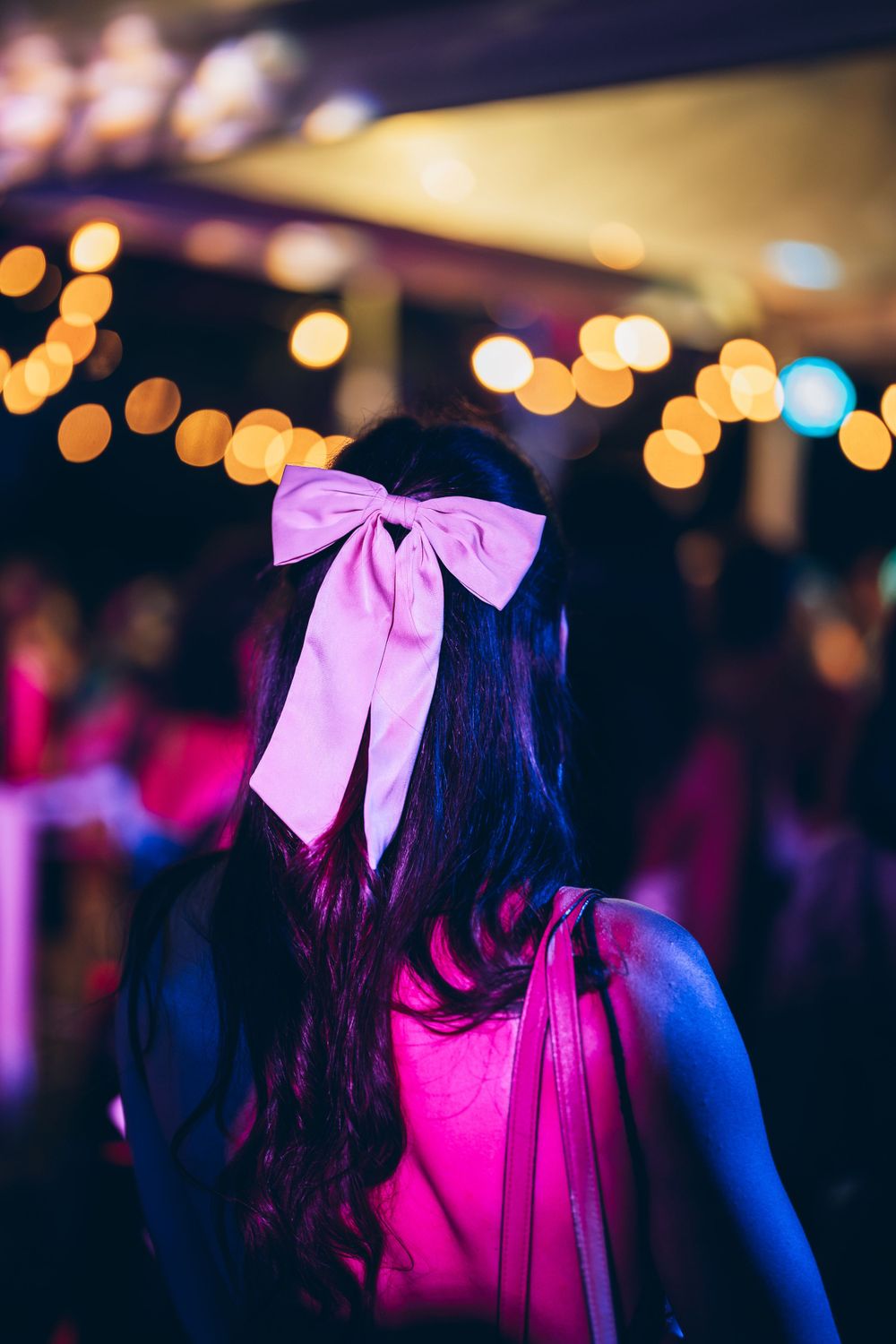 Girl with long dark hair wearing pink bow in nightclub with bokeh lights.