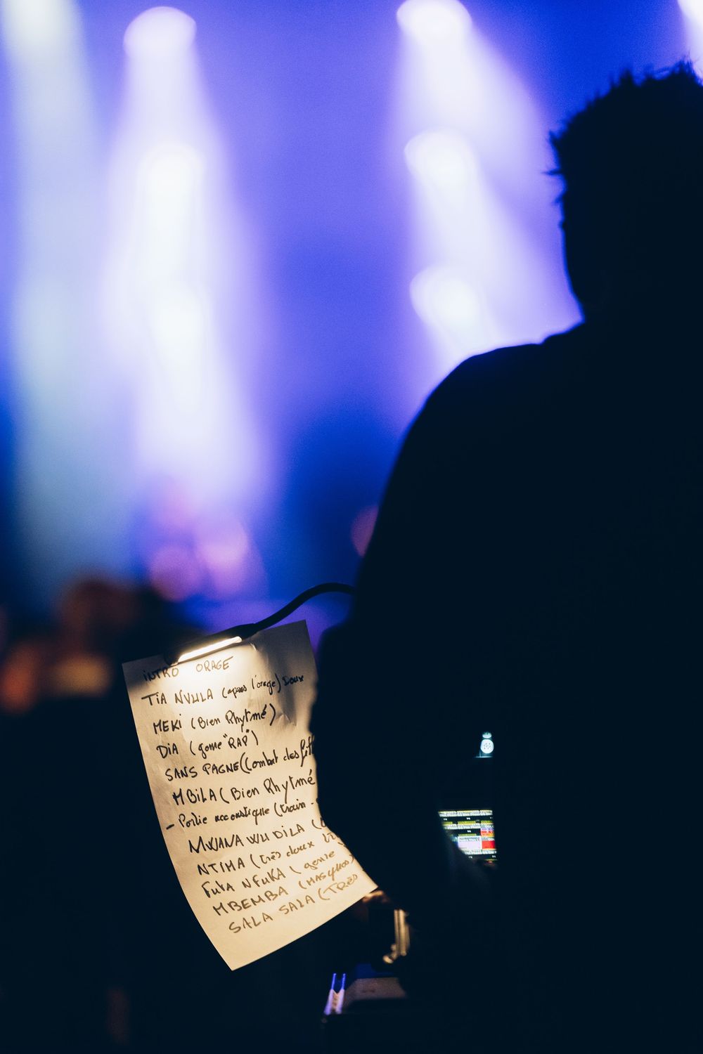 Silhouetted figure holds a handwritten setlist at a live concert with blue stage lights glowing in the background.