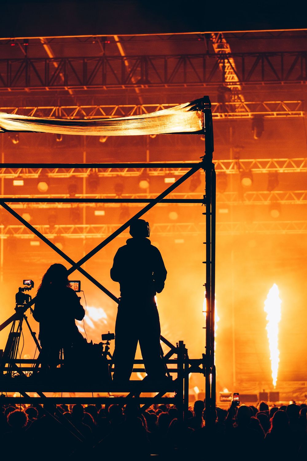 Silhouettes of camera crew on scaffolding at a live concert with dramatic orange pyrotechnic flames and stage lighting.