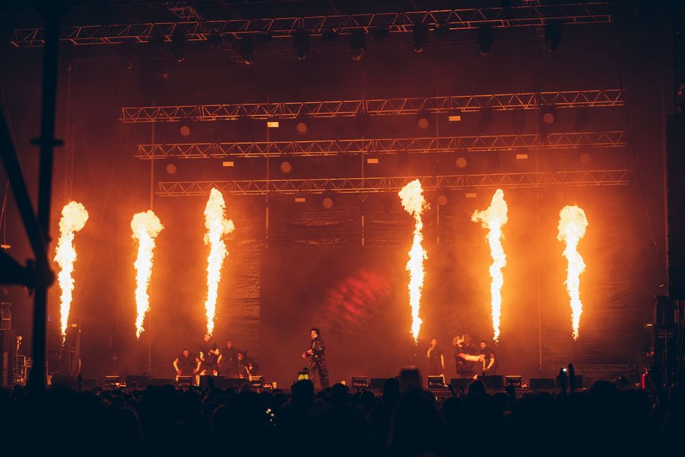 Live concert stage with massive fire pyrotechnics shooting flames into the dark sky above a crowd of silhouetted fans.