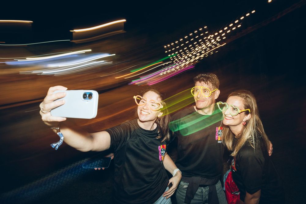Four friends take a nighttime selfie with glowing glasses, colorful light trails streaking behind them.
