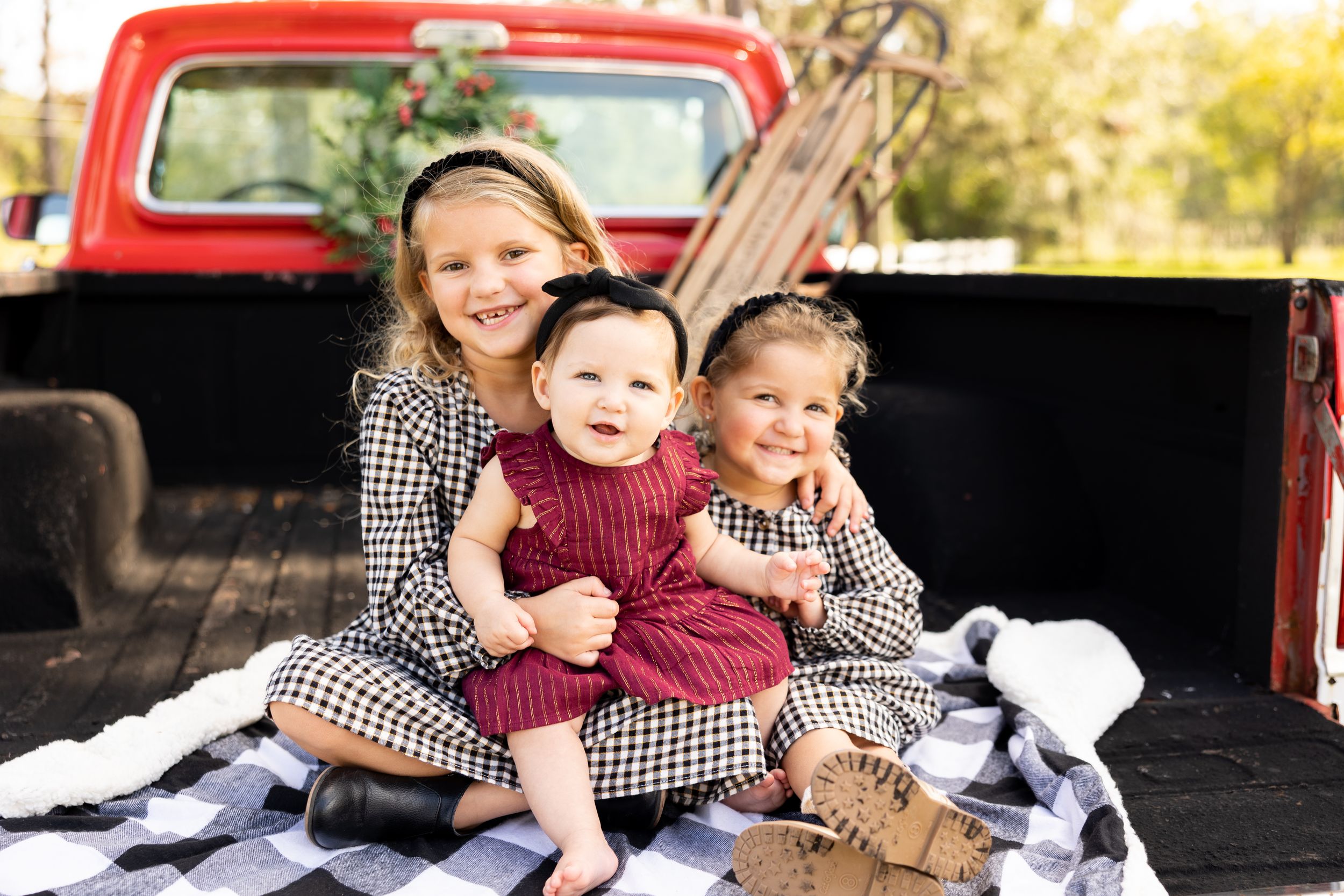 Three young sisters in matching outfits sit smiling on a red truck bed with a plaid blanket during a fall photo shoot.