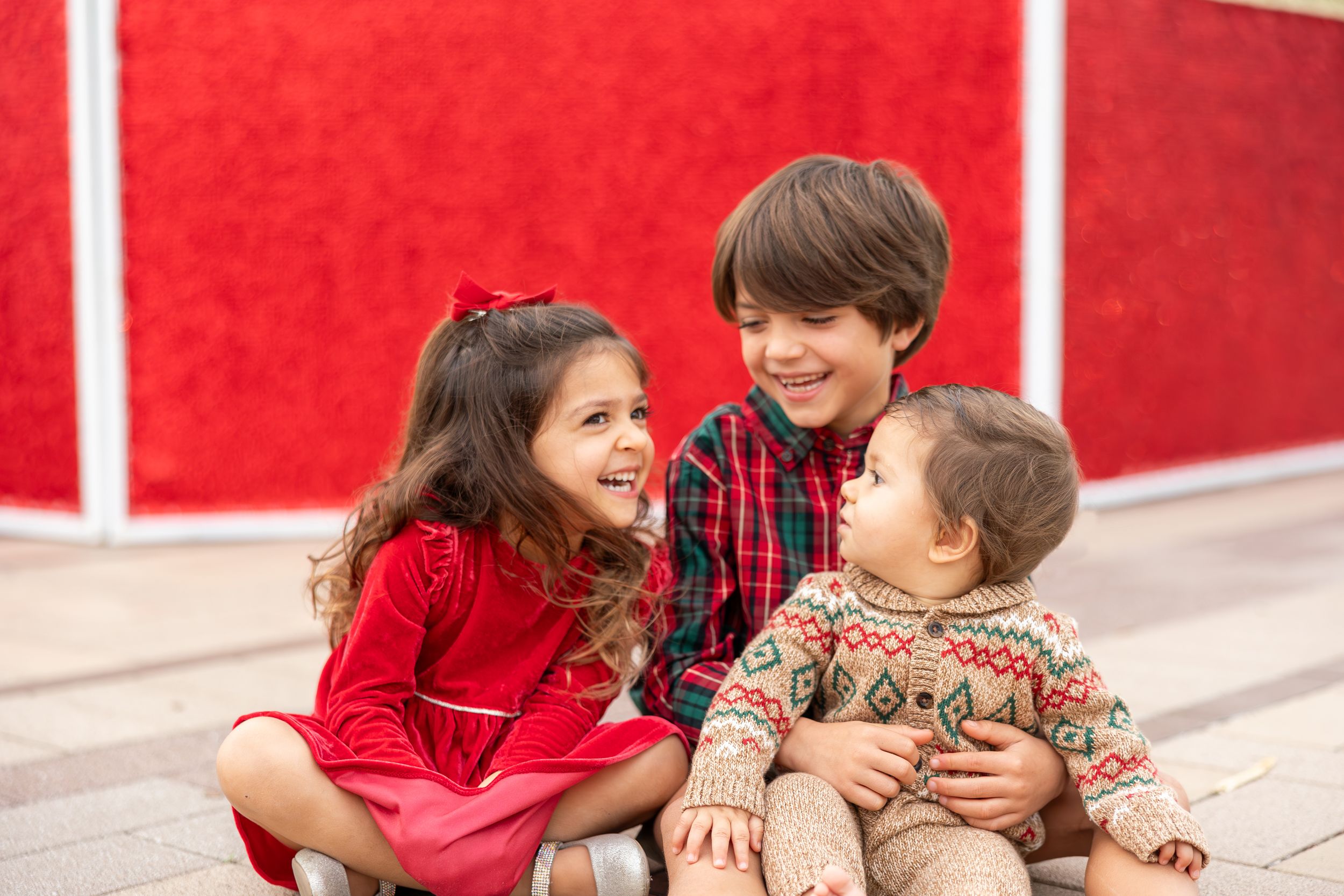 Three happy children in festive outfits sit together smiling in front of a bright red background.
