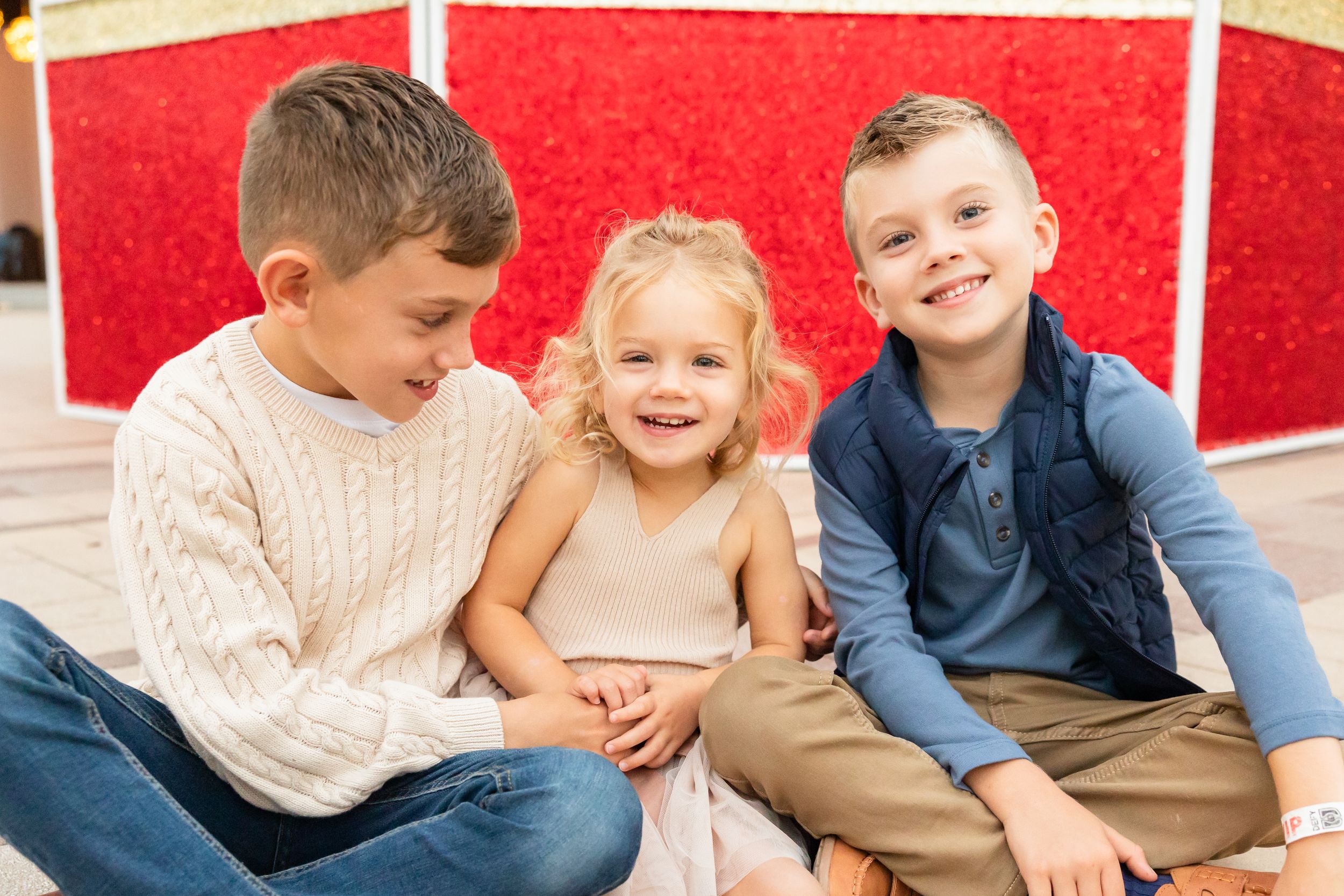 Three happy children sitting together, two boys and a girl, posing in front of a red floral backdrop.