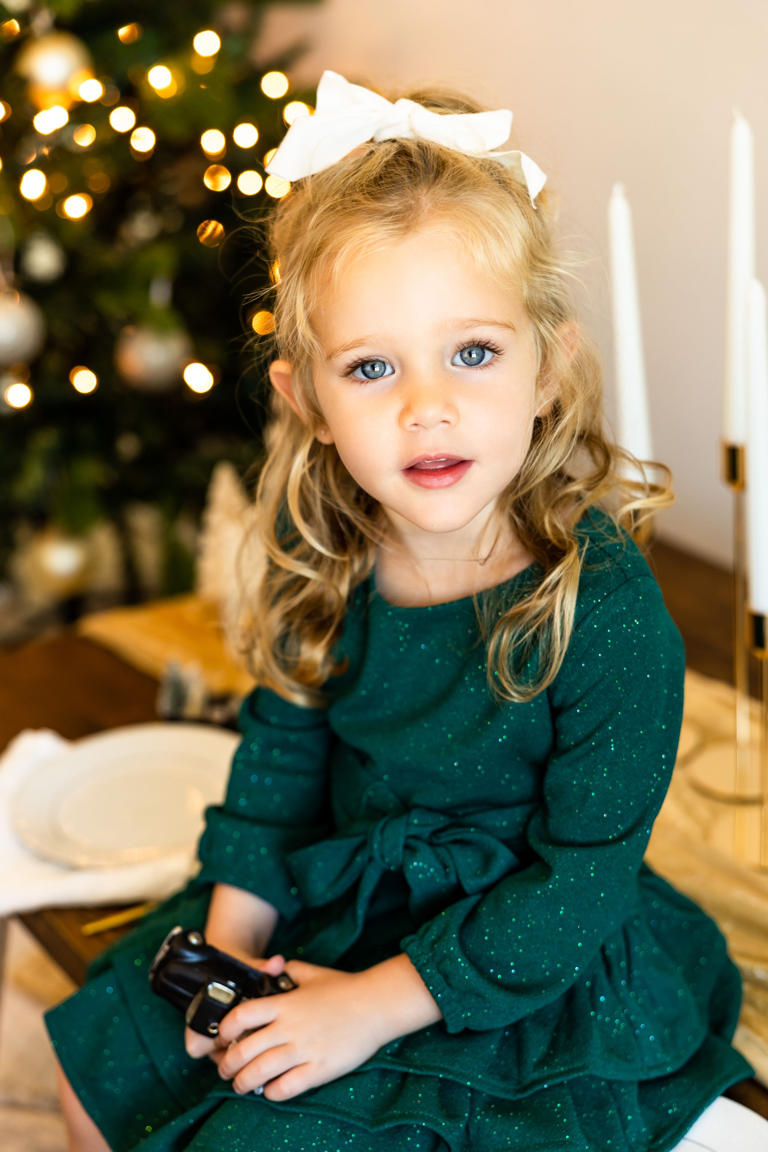 Young girl in green sparkle dress with white bow sits near Christmas tree holding a toy car.