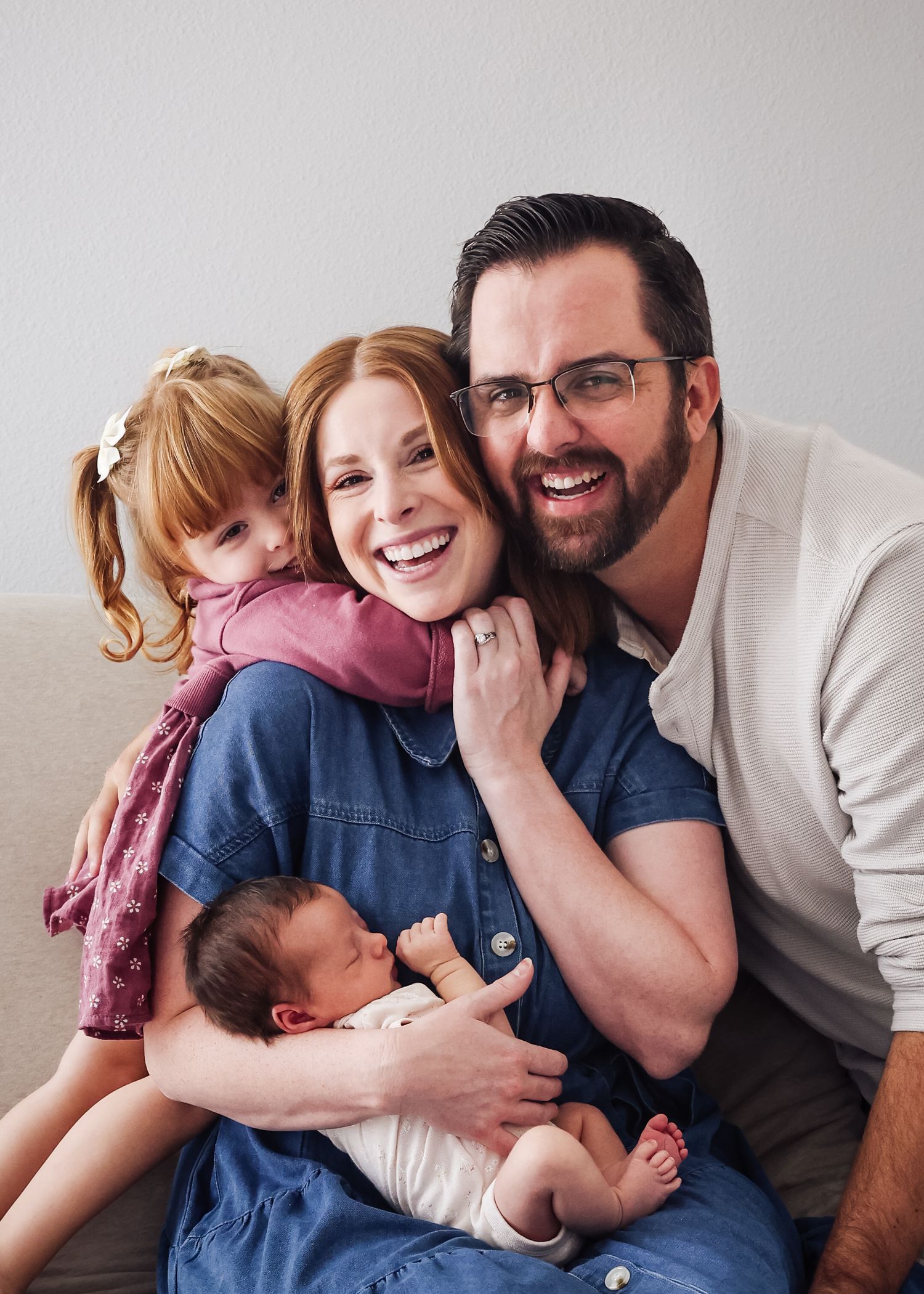 Happy family portrait with smiling parents holding newborn baby and young toddler girl on a cozy couch.