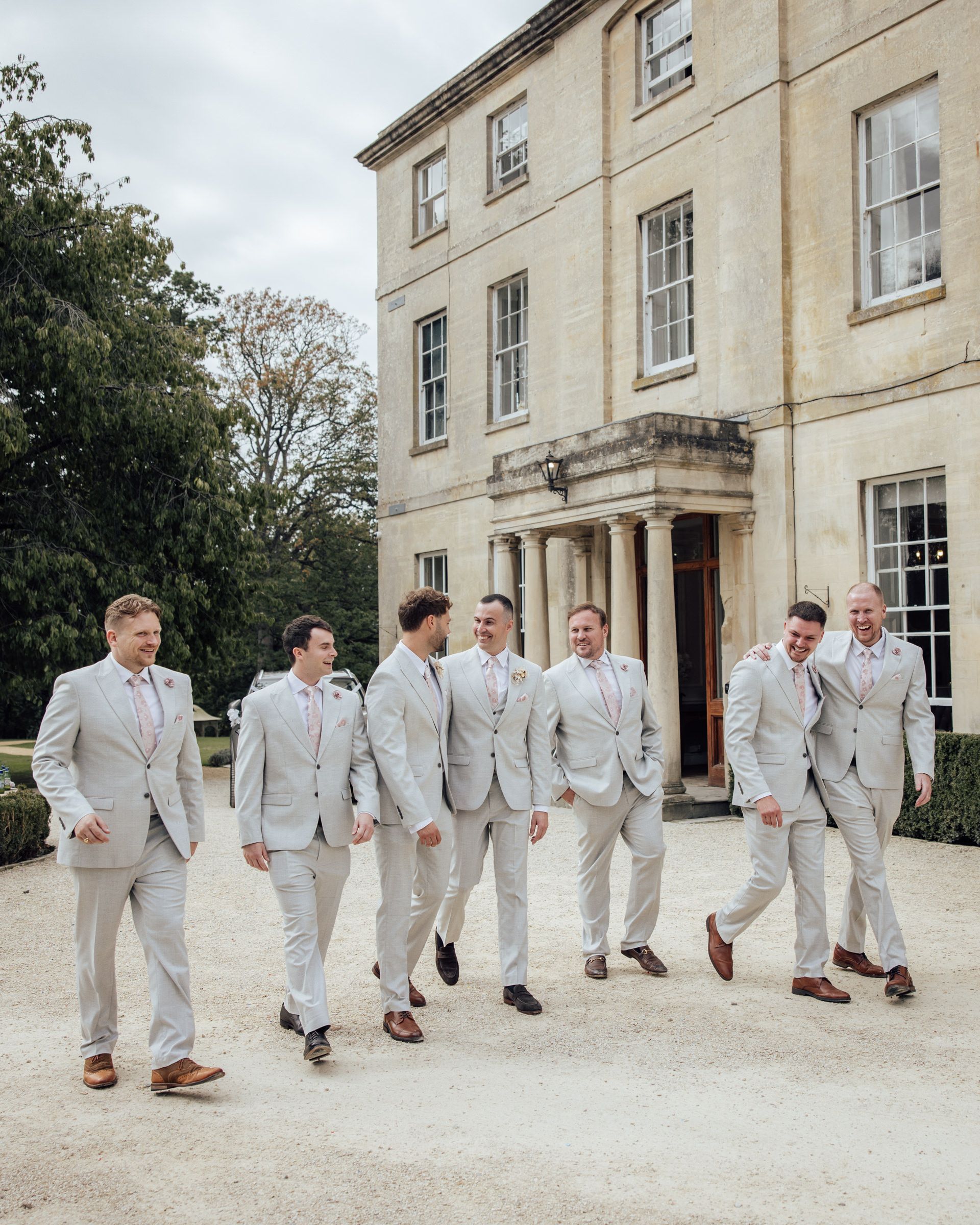 Seven groomsmen in matching light gray suits walk together on a gravel path in front of an elegant stone manor house.
