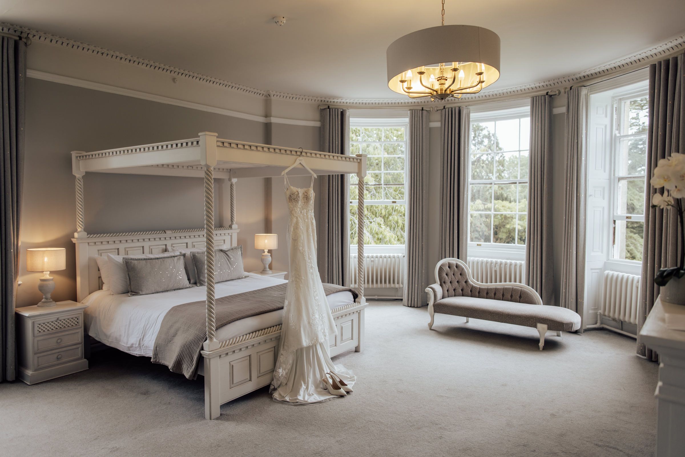 Elegant bridal suite with a white four-poster bed, a wedding dress hanging from the canopy, tall windows, a chaise lounge and a chandelier.