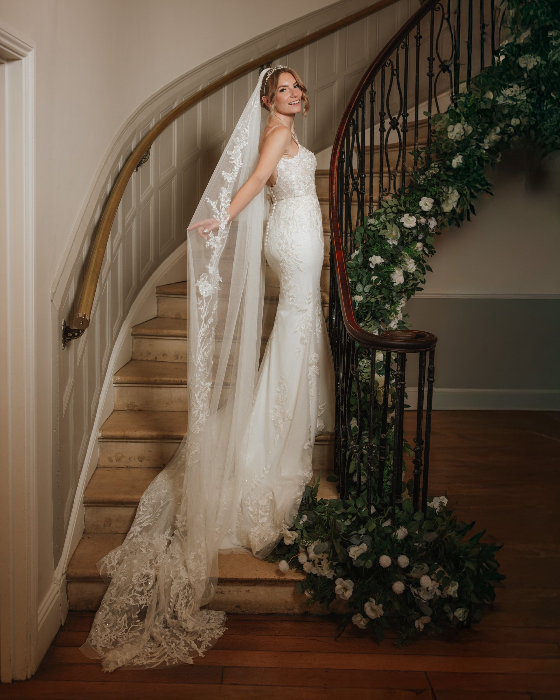 Bride in a lace wedding gown and long cathedral veil poses on a curved wooden staircase draped with white floral garland.