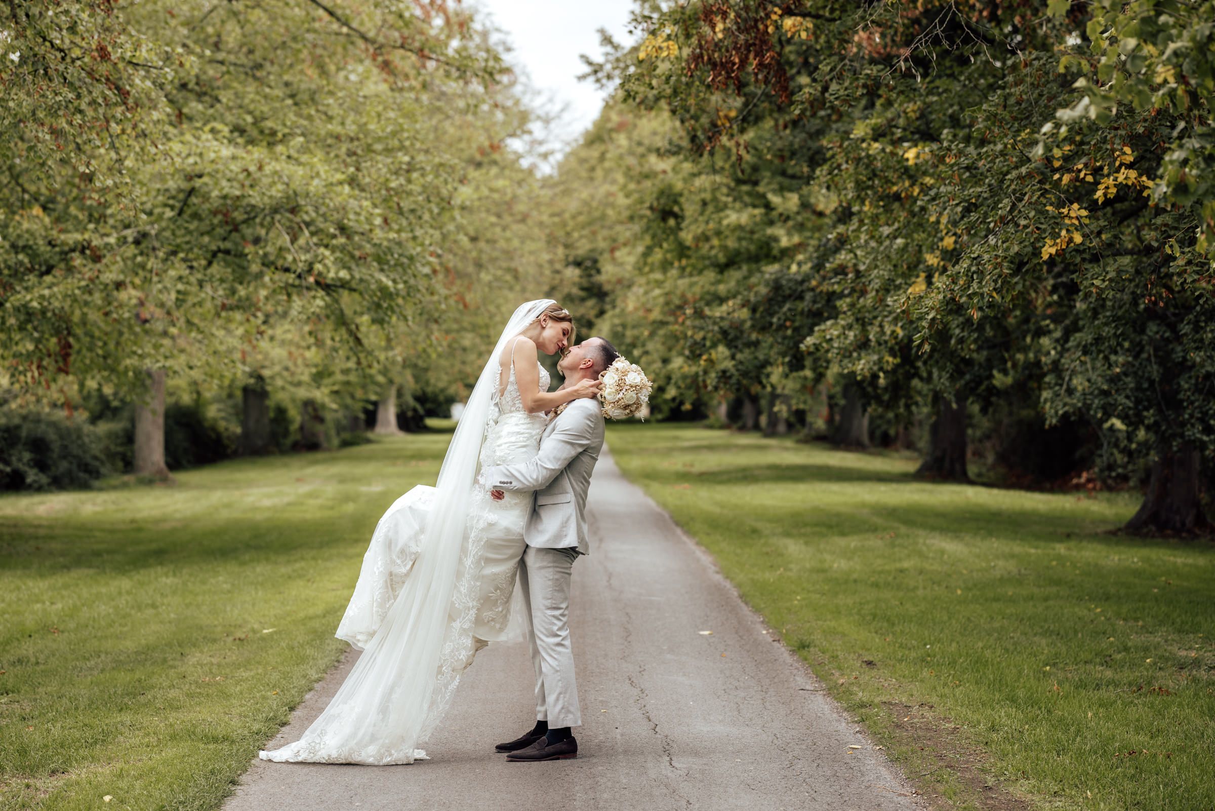 Groom in a light suit lifts his bride in a flowing white wedding gown while kissing her on a tree-lined path in a sunny park.