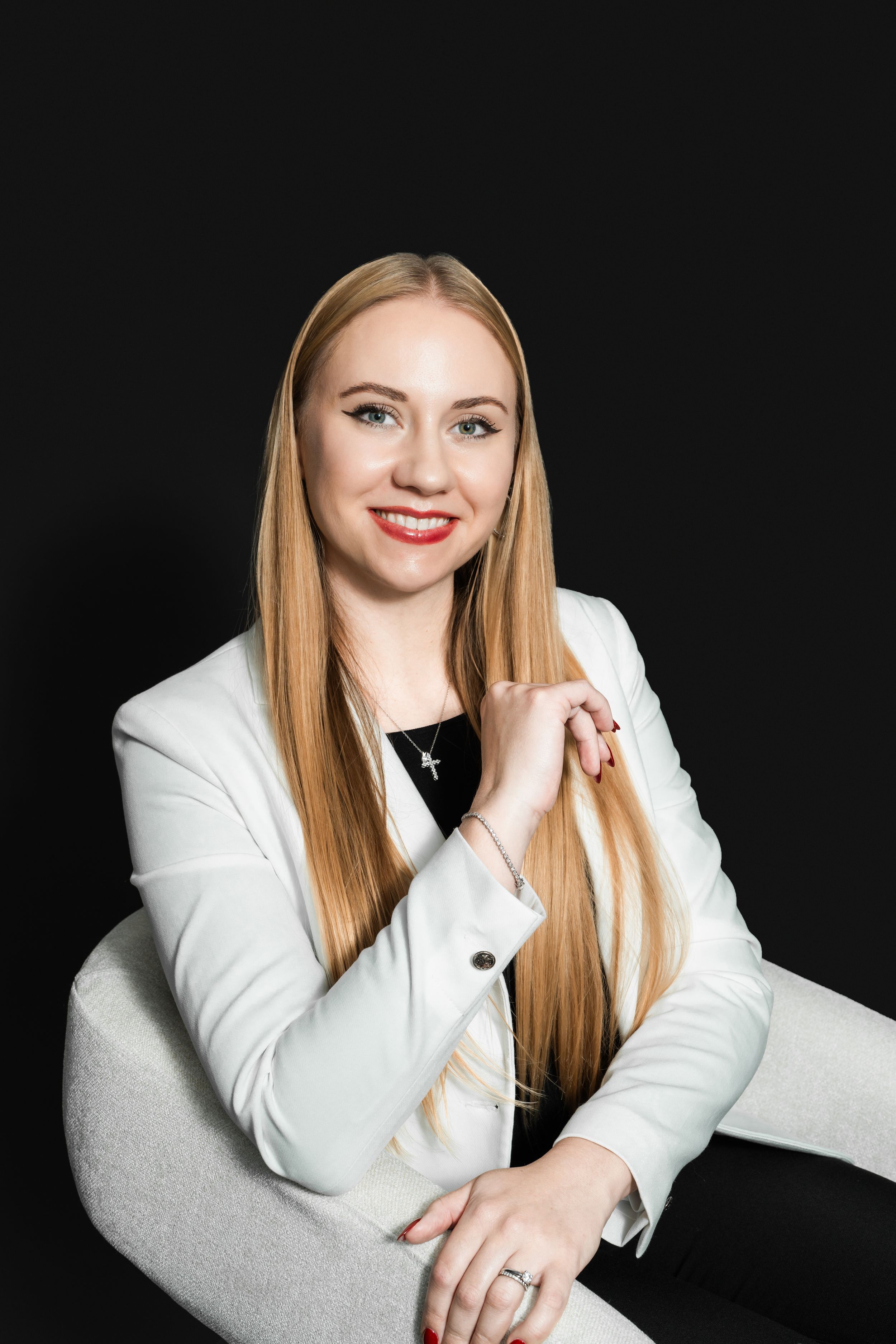 Smiling woman with long blonde hair and red lipstick wearing a white blazer, seated in a cream chair against a black background.