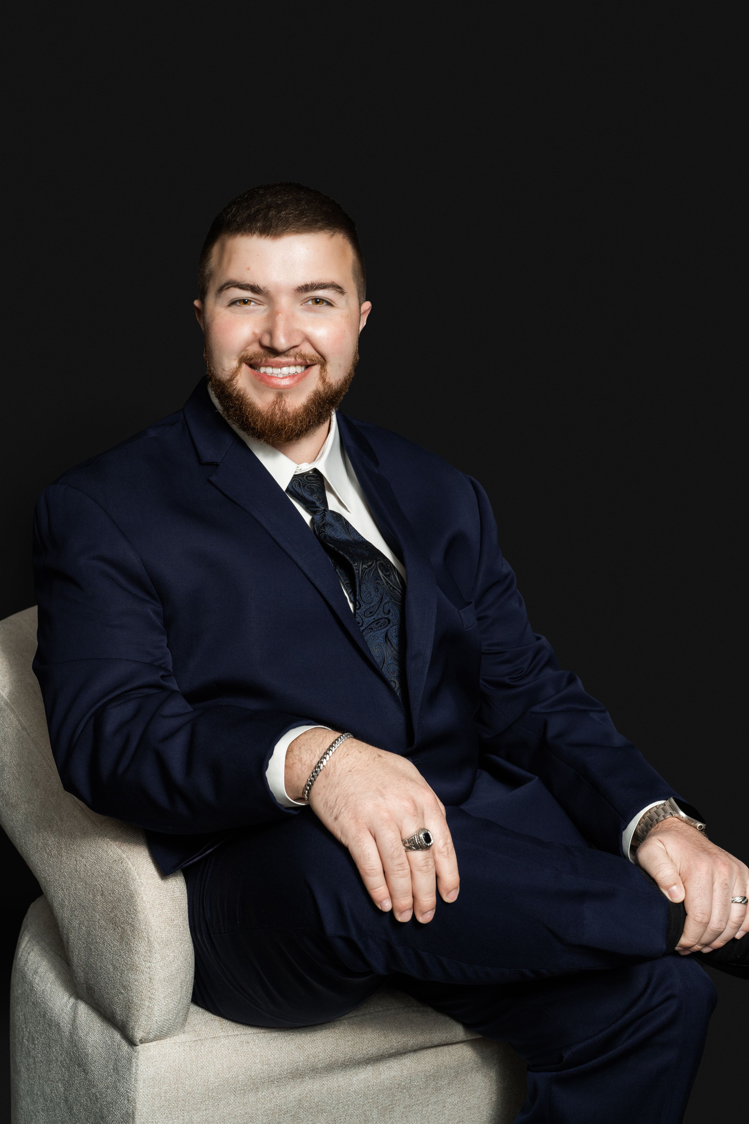 Smiling bearded man in a navy suit and patterned tie sits in a light upholstered chair against a dark studio background.