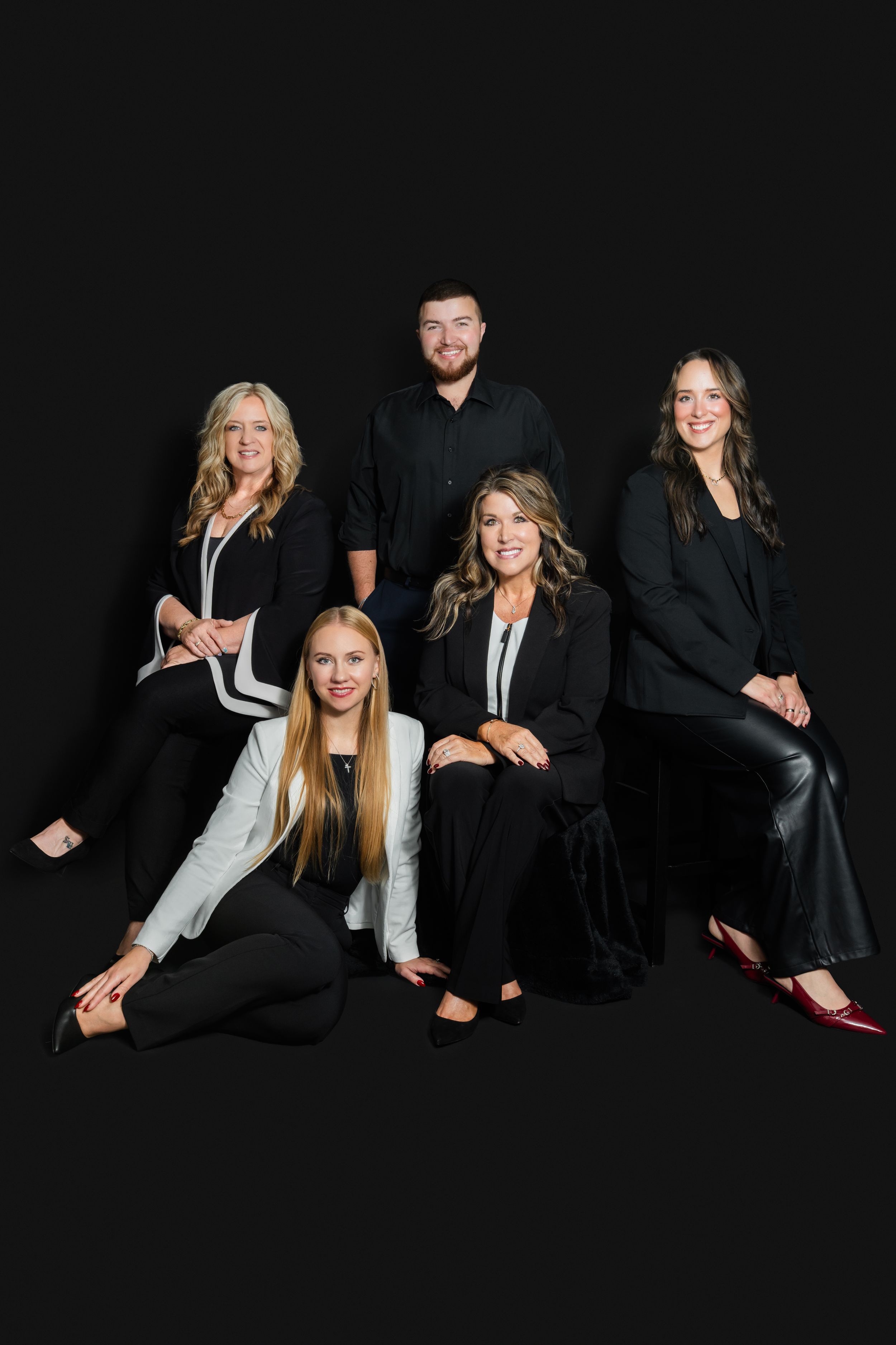 Professional group portrait of four women and one man in coordinated black attire posed against a dark studio background.