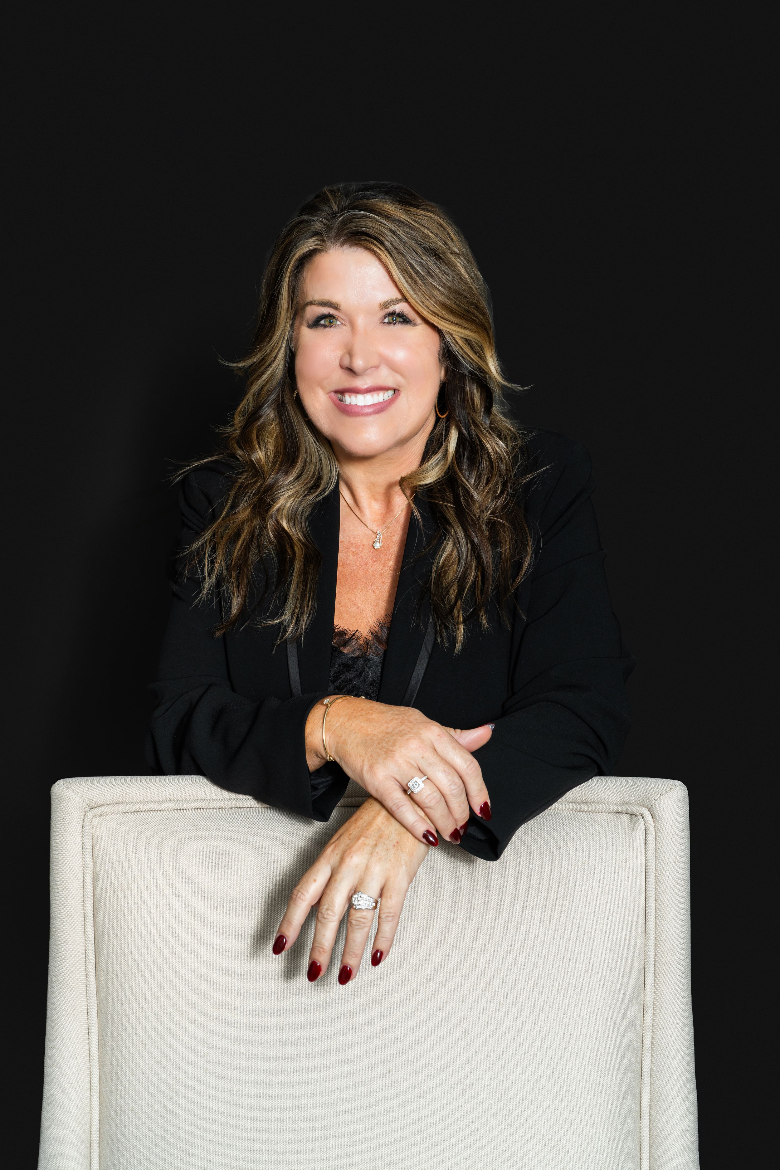 Smiling woman with long wavy hair in a black blazer leans on the back of a cream upholstered chair against a dark background.