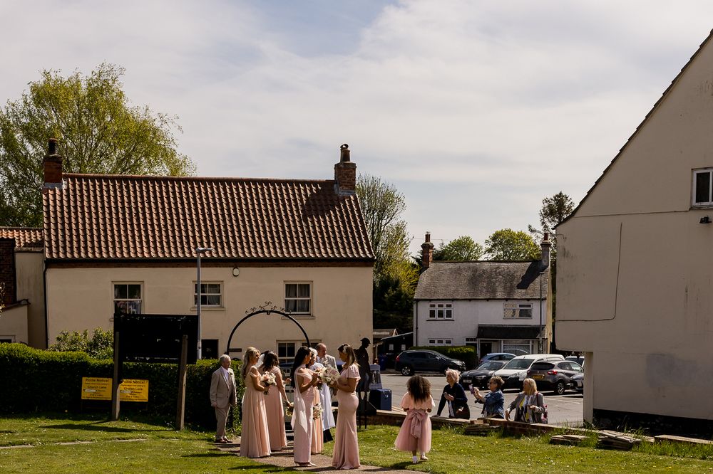 Wedding party in pink dresses gathered outdoors near a rustic English countryside venue on a sunny day.