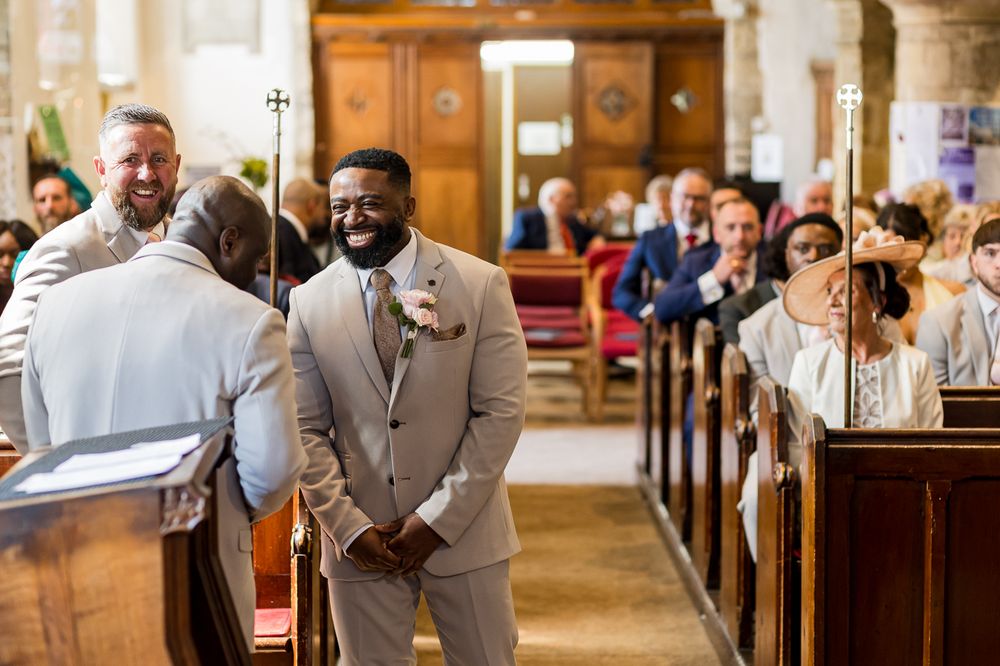Groom in a light gray suit smiling at the altar during a church wedding ceremony with guests seated in pews.