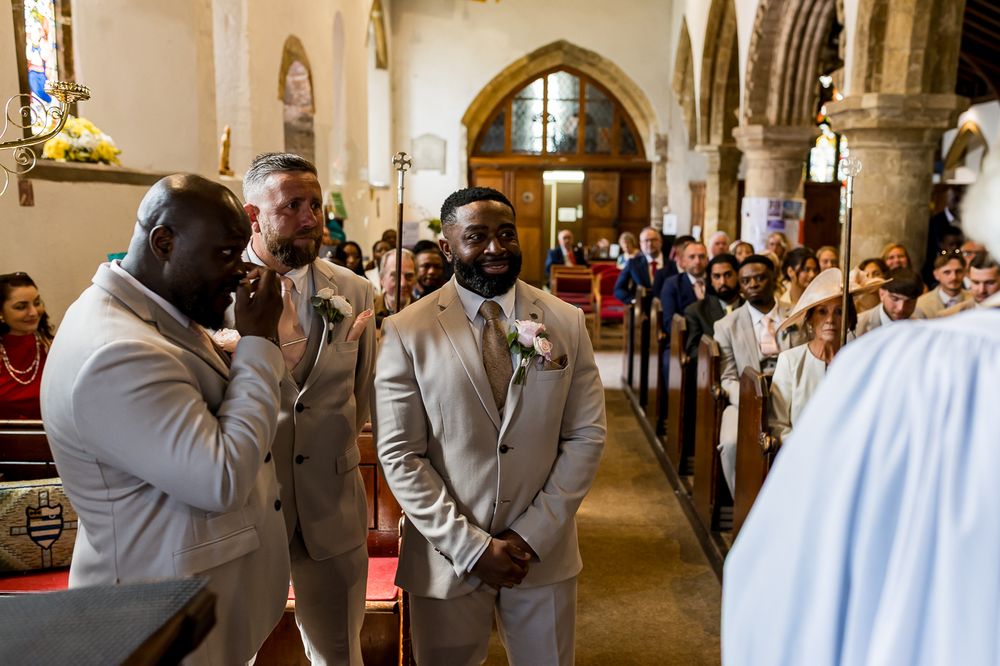 Groom and groomsmen in tan suits stand at church altar during wedding ceremony, with guests seated in pews behind them.