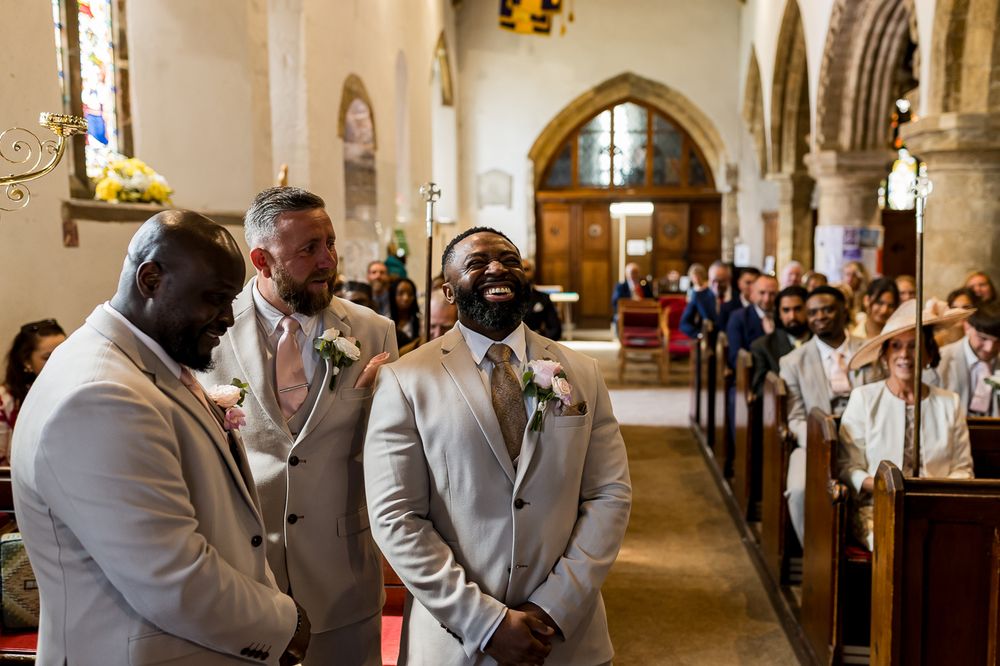 Groom smiling at the altar with groomsmen in beige suits inside a traditional church on his wedding day.