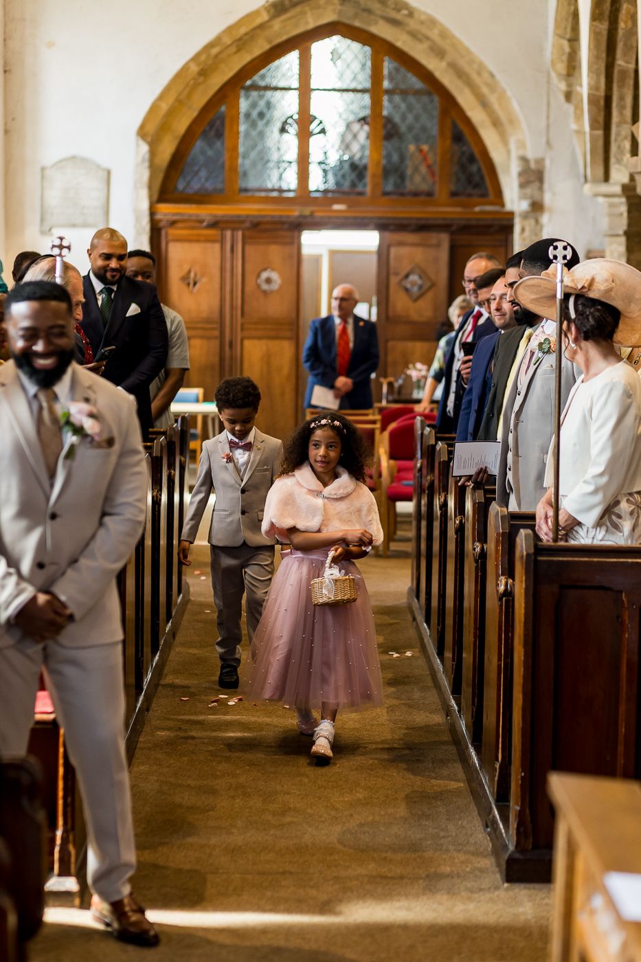 Flower girl and page boy walk down church aisle at wedding ceremony, guests standing in wooden pews.