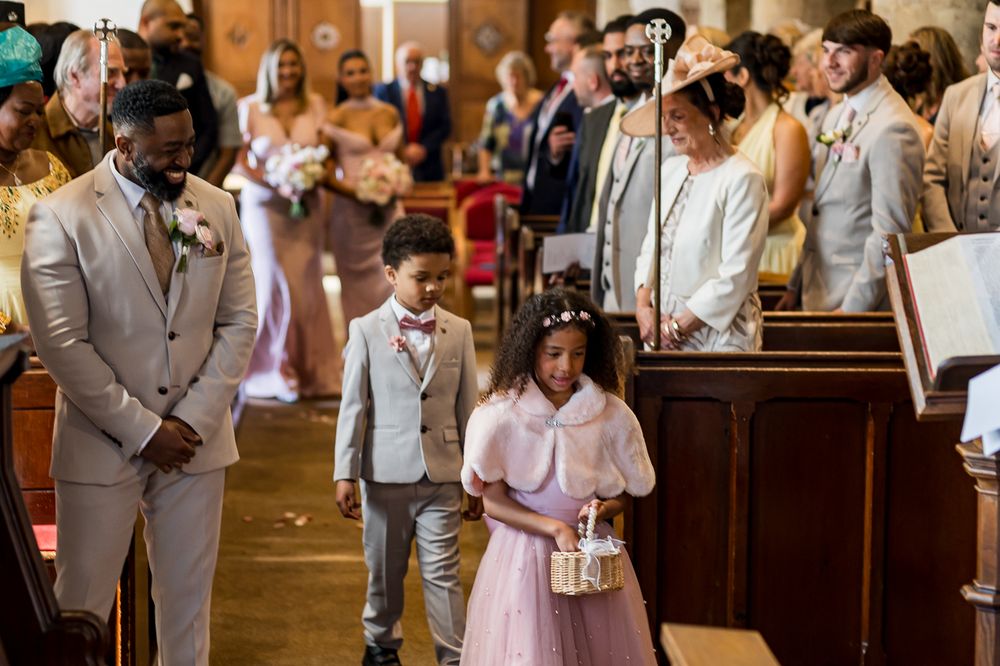 Children in formal attire walk down a church aisle during a wedding ceremony, with guests seated in pews.