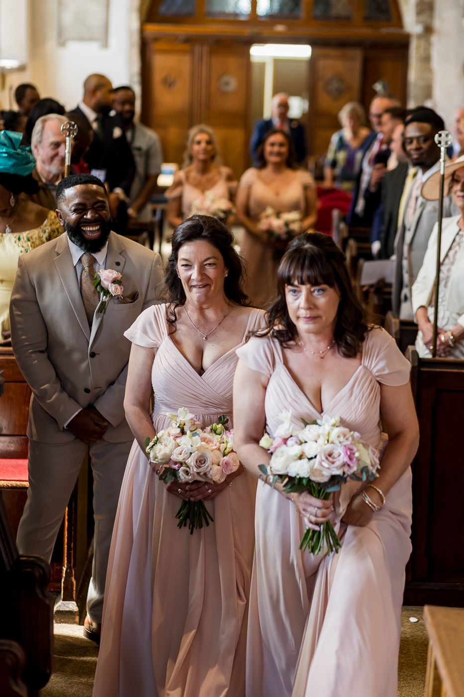 Two bridesmaids in blush gowns walk down a church aisle holding floral bouquets at a wedding ceremony.