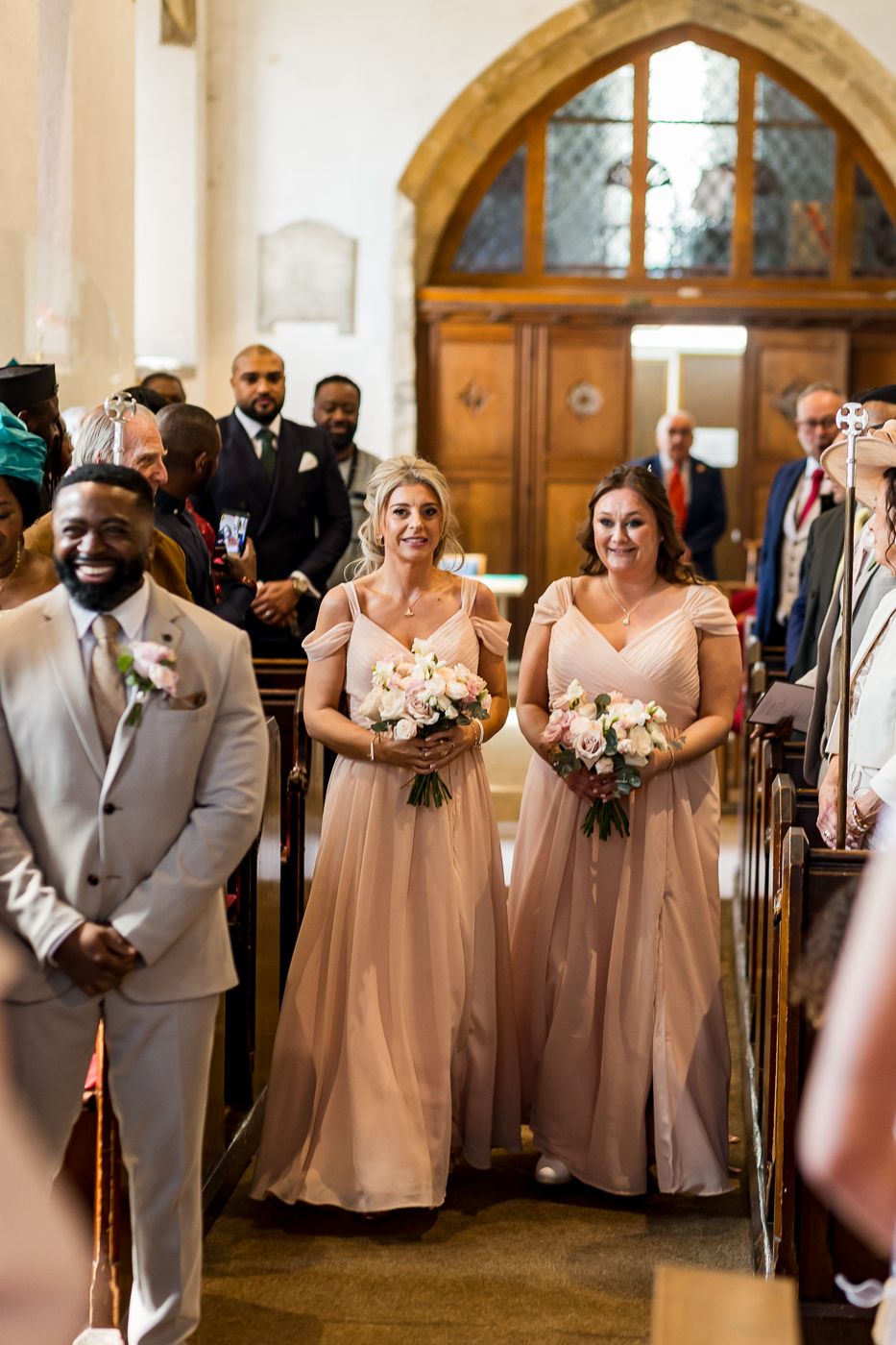 Two bridesmaids in blush pink gowns walk down a church aisle holding floral bouquets during a wedding ceremony.