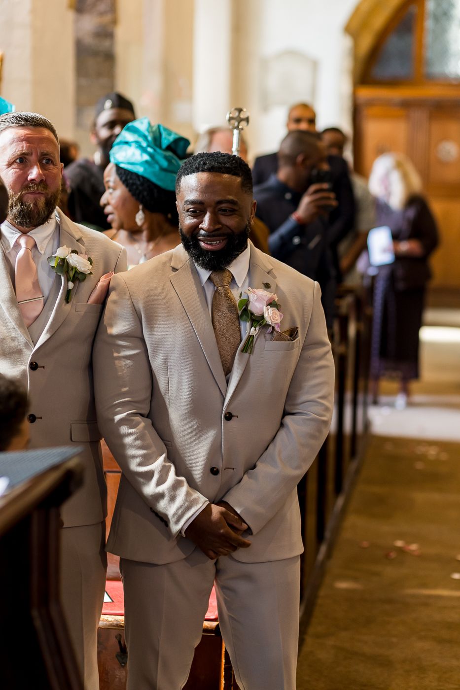 Groom in a tan suit smiling at the altar during a church wedding ceremony with guests in the background.