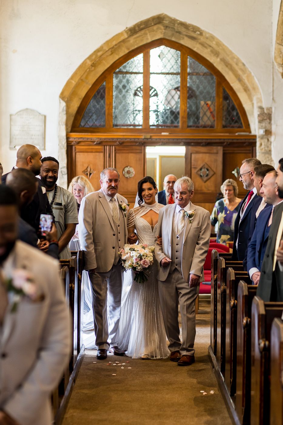 Bride walks down church aisle escorted by two men in suits, holding bouquet, guests seated in pews of stone chapel.