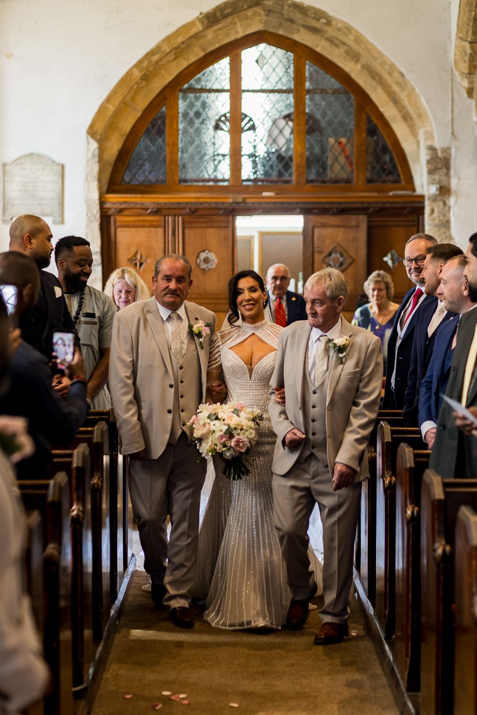 Bride walks down church aisle escorted by two men in tan suits, holding a bouquet, with guests watching.