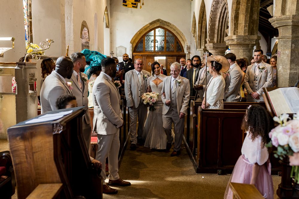 Bride walks down the aisle in a church wedding ceremony, surrounded by groomsmen in gray suits.