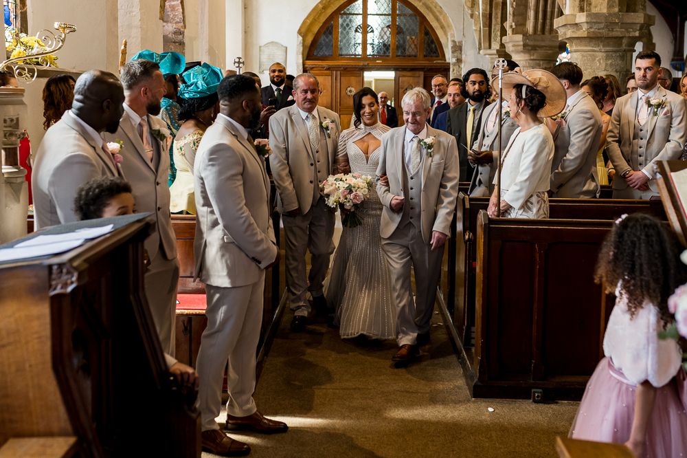 Bride walks down church aisle with escort past groomsmen in grey suits during elegant wedding ceremony.