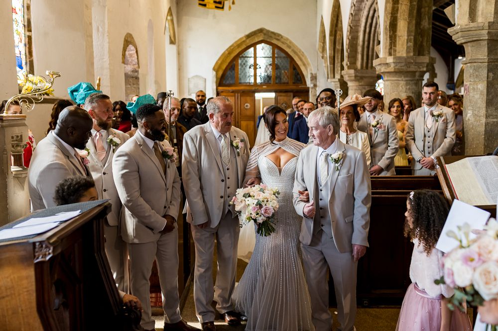 Bride walks down the aisle in a church wedding ceremony surrounded by groomsmen in beige suits.