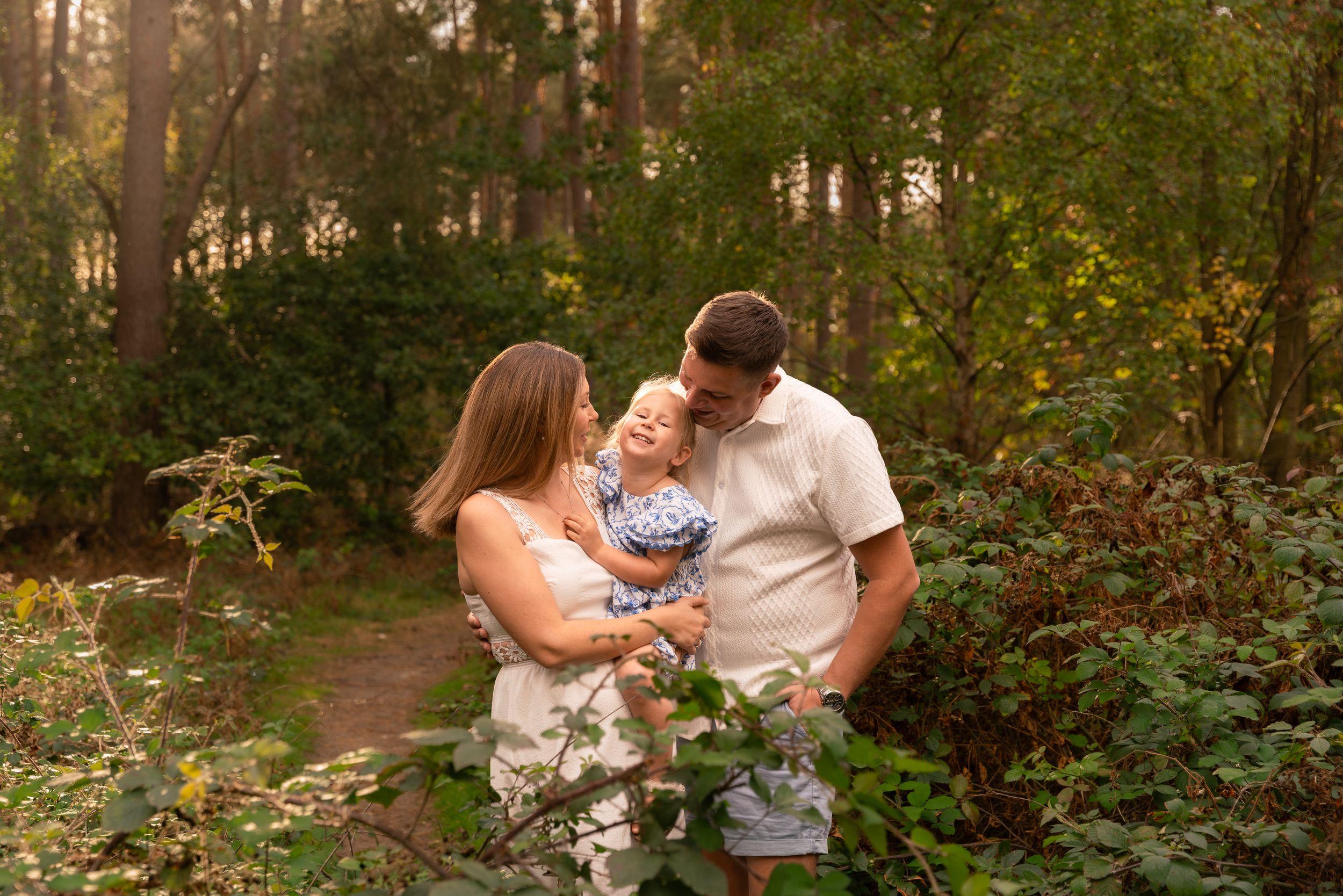 Family of three sharing a tender moment on a forest path surrounded by lush green foliage.