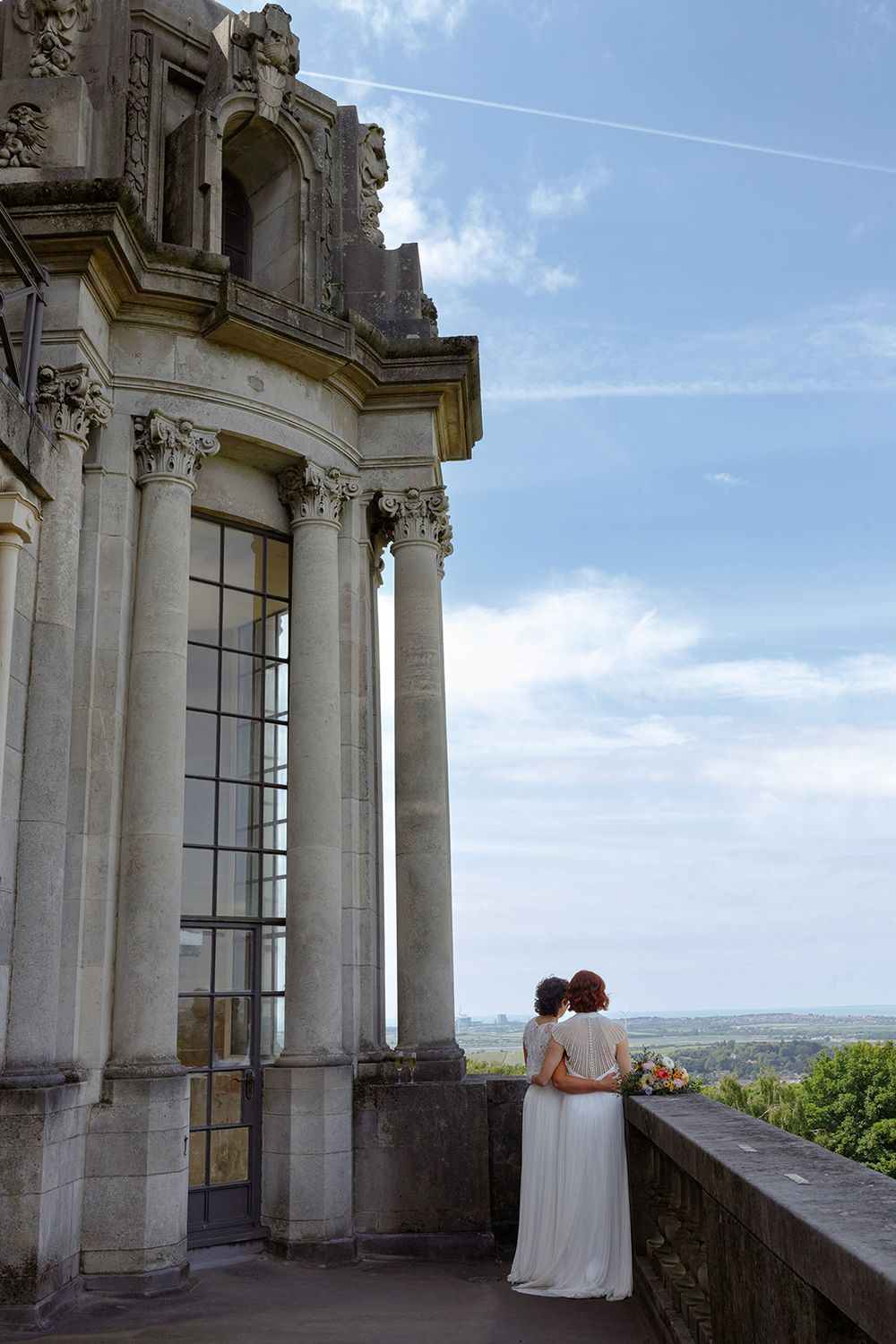 Two brides in white dresses embrace on a castle balcony overlooking scenic countryside under a blue sky.