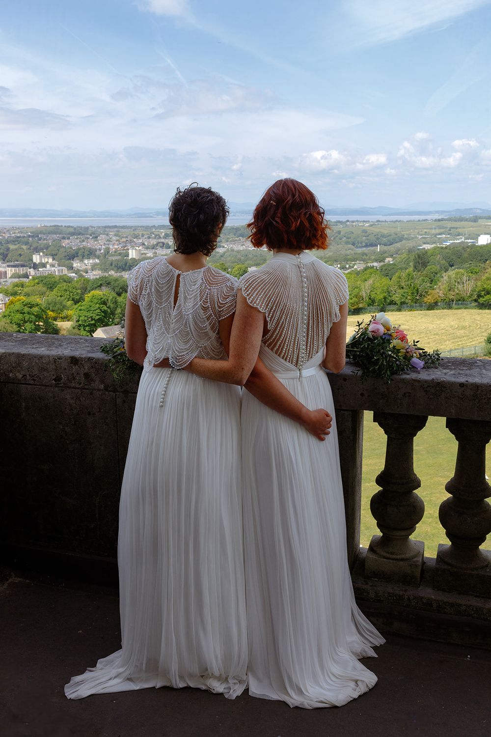 Two brides in white dresses stand on a stone balcony overlooking a scenic landscape on their wedding day.