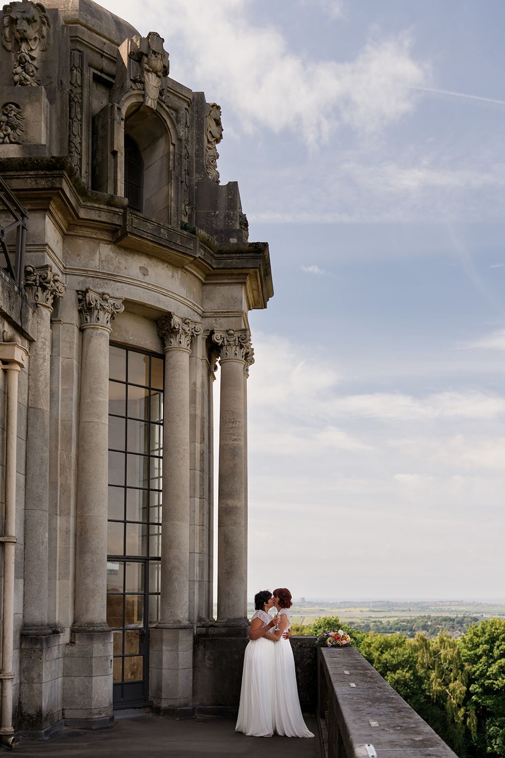 Two brides in white dresses share a romantic moment on a grand baroque palace balcony overlooking a scenic landscape.